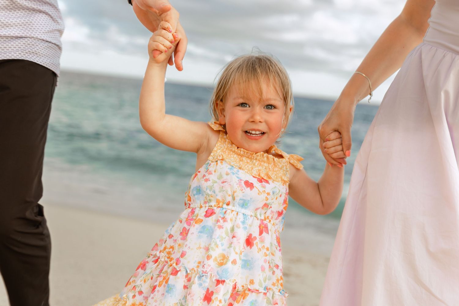 A toddler in a floral dress walks along the beach while holding hands with an adult in white clothing.