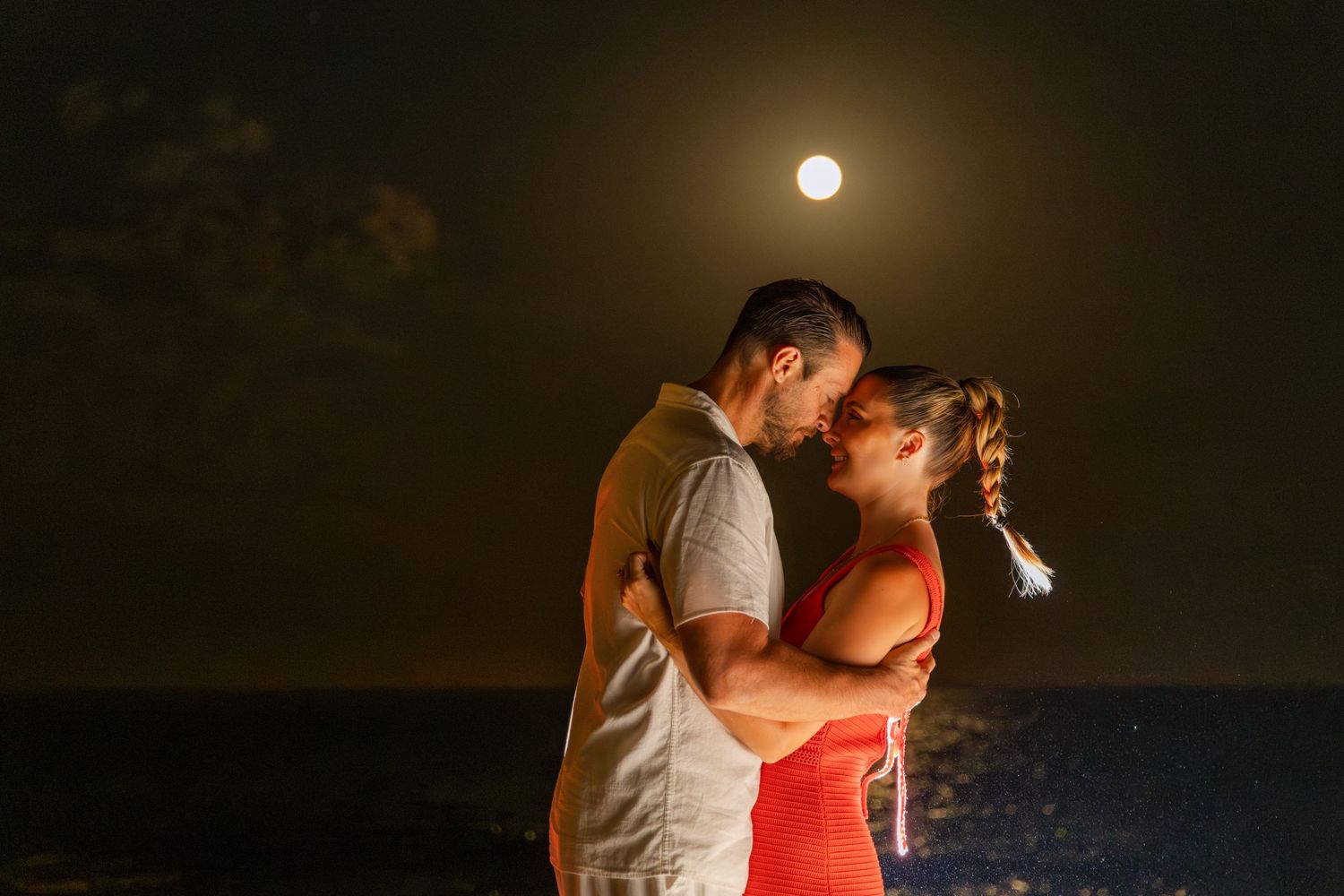 A silhouetted couple embraces against a dramatic moonlit sky, creating a romantic nighttime scene.