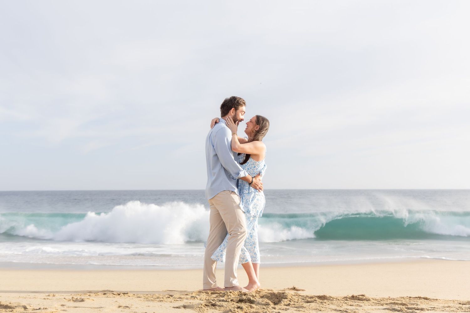 A couple in white outfits embraces on a scenic beach with crashing waves and golden sand at sunset.
