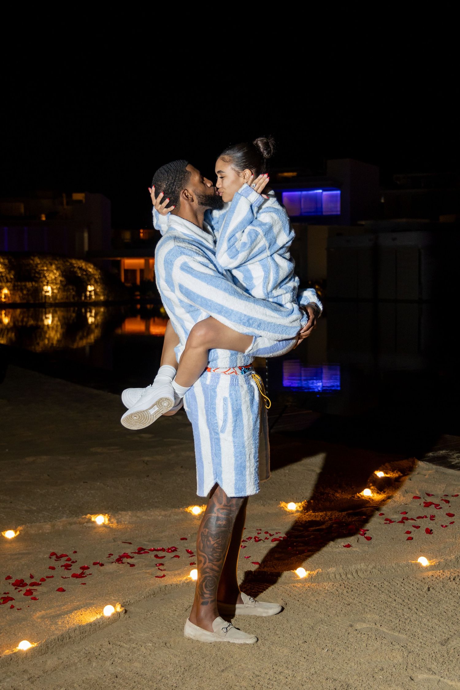 Couple in matching blue and white striped outfits embrace at night with candles and rose petals on the beach.