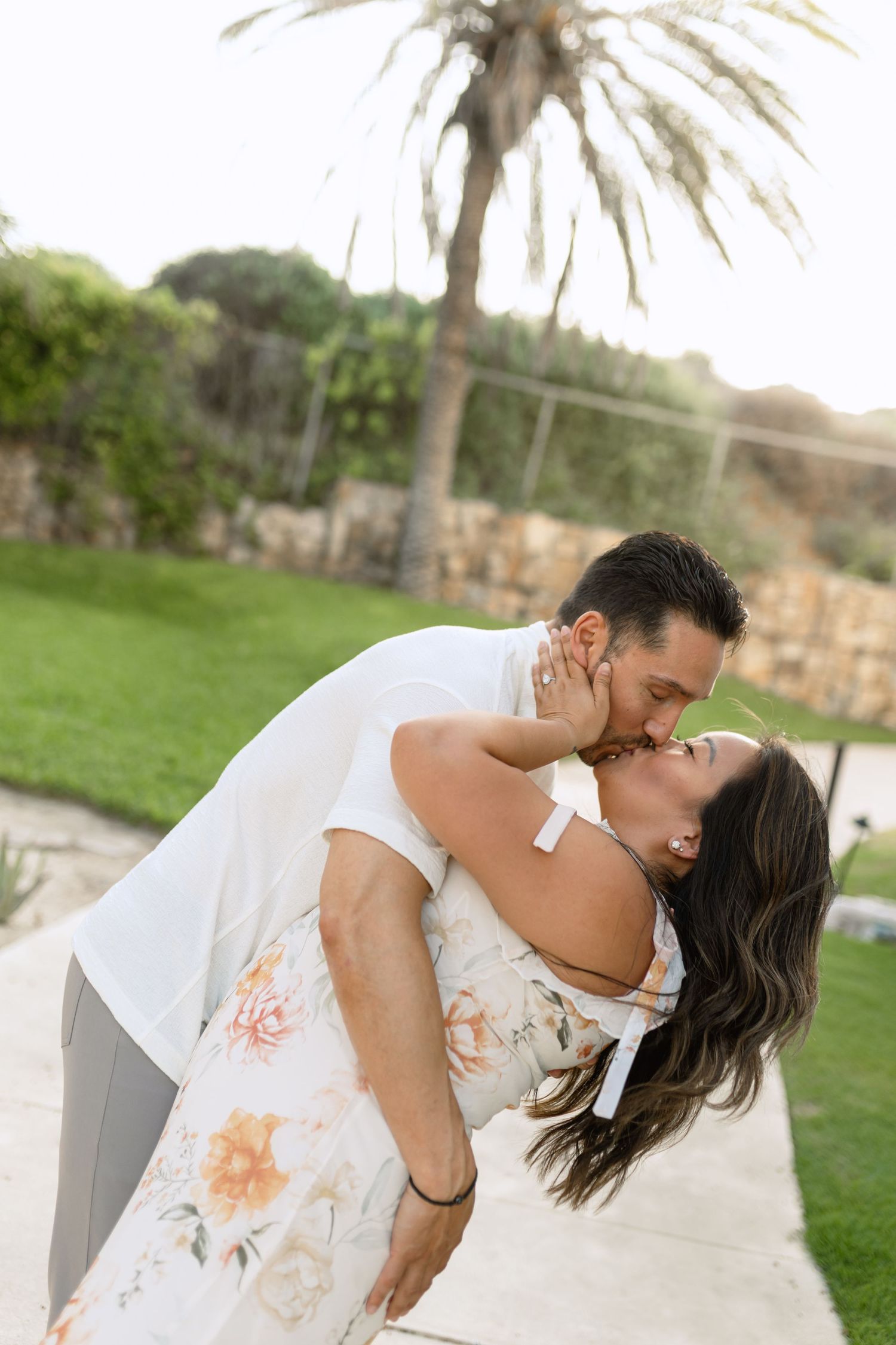 A romantic couple in white attire share an intimate dance pose on a grassy lawn with palm trees in the background.
