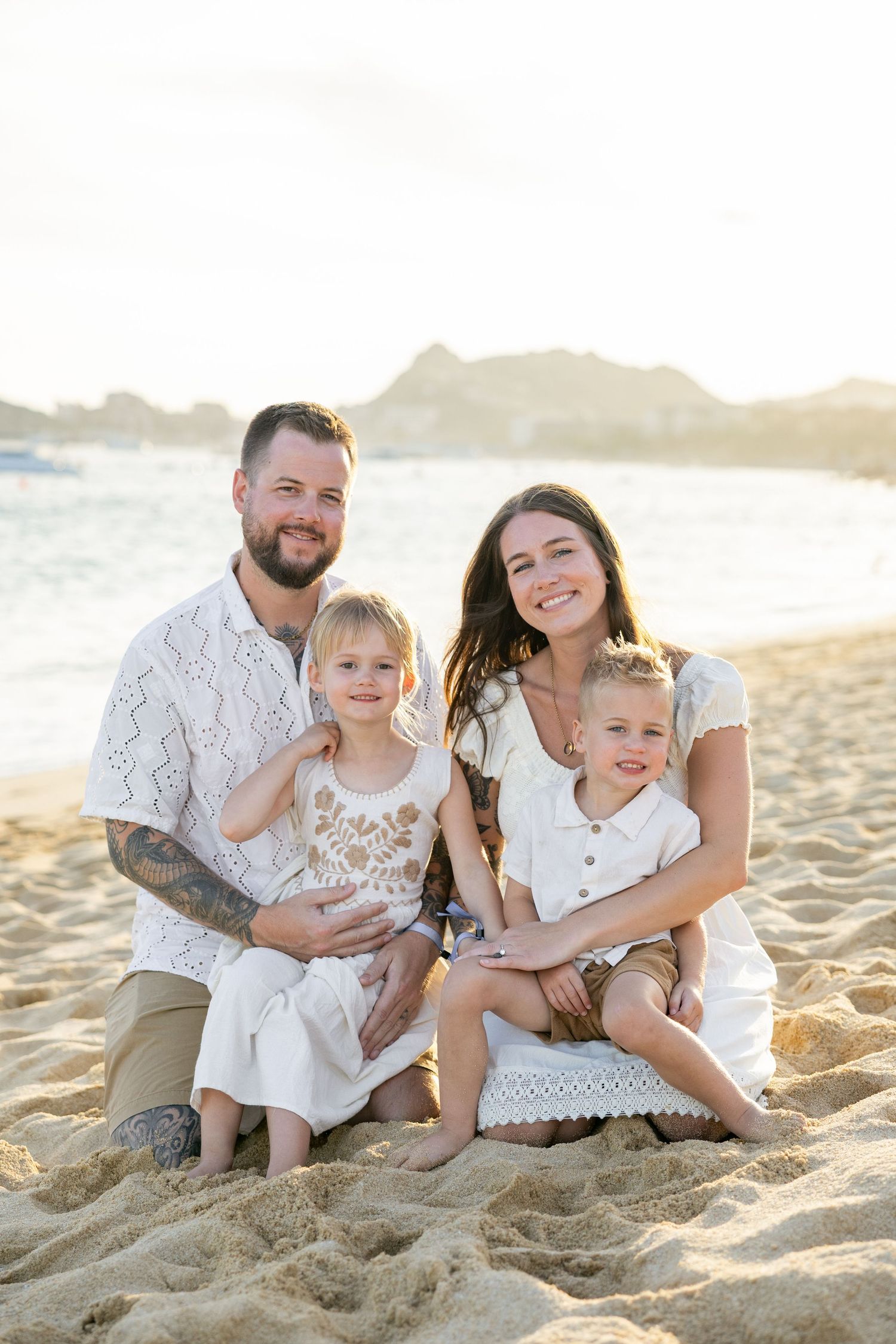 Family wearing white outfits sits together on sandy beach during golden hour sunset in medano beach los cabos