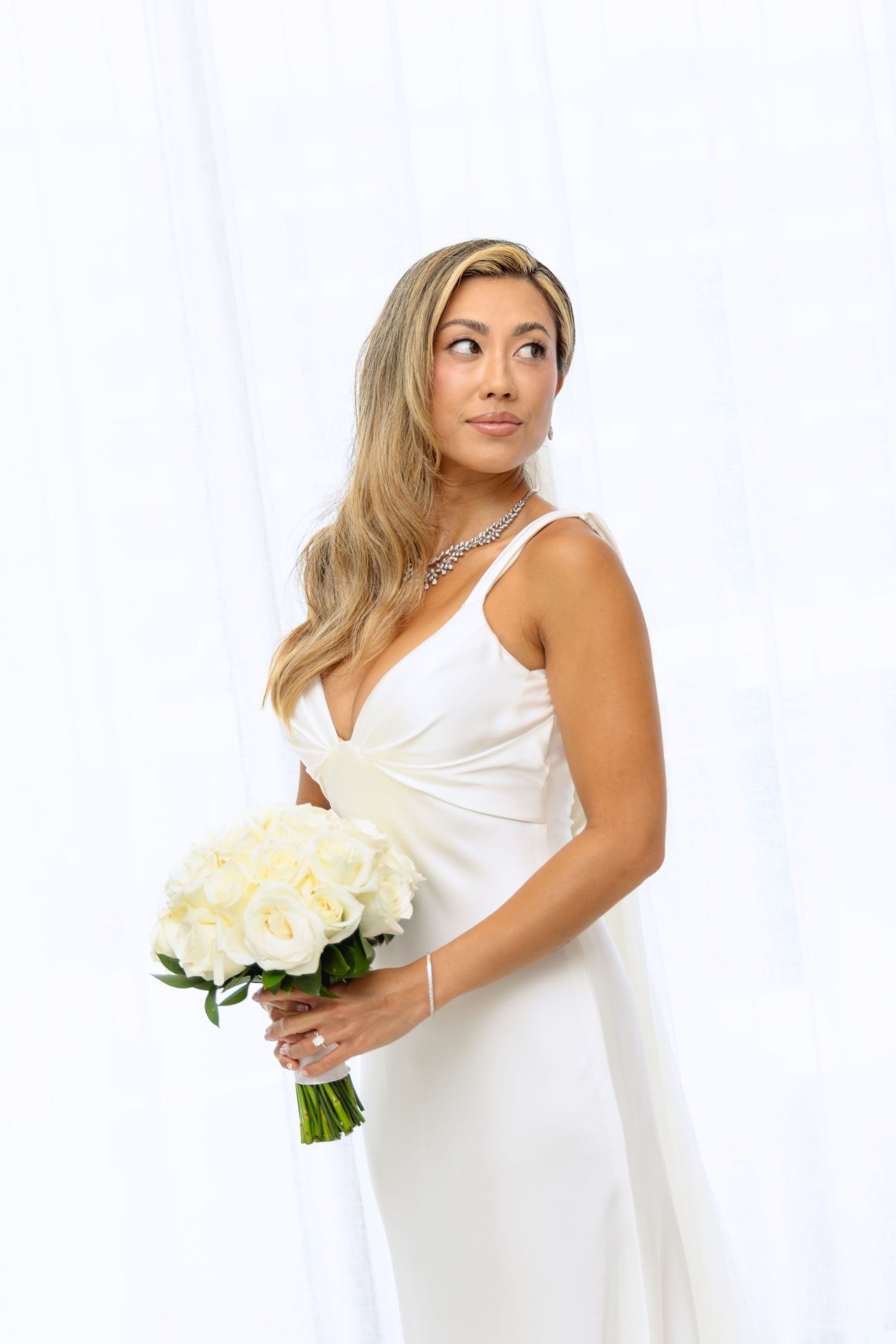 Bride in sleek white dress holds white roses while posing playfully.