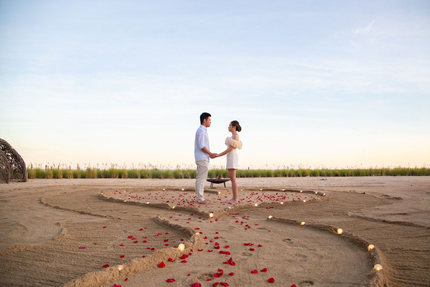 Beach proposal setup with rose petals and candles arranged on sandy shore.