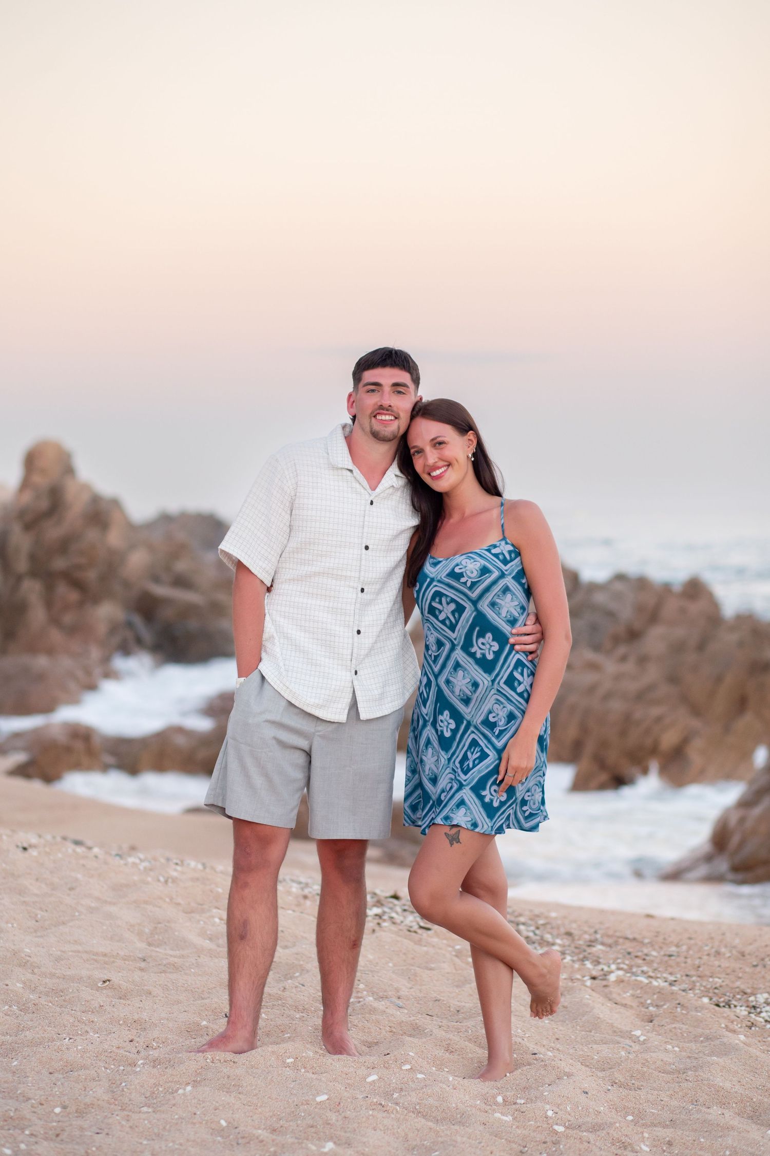 A couple in casual beach attire shares a playful moment together on a sandy beach at sunset.