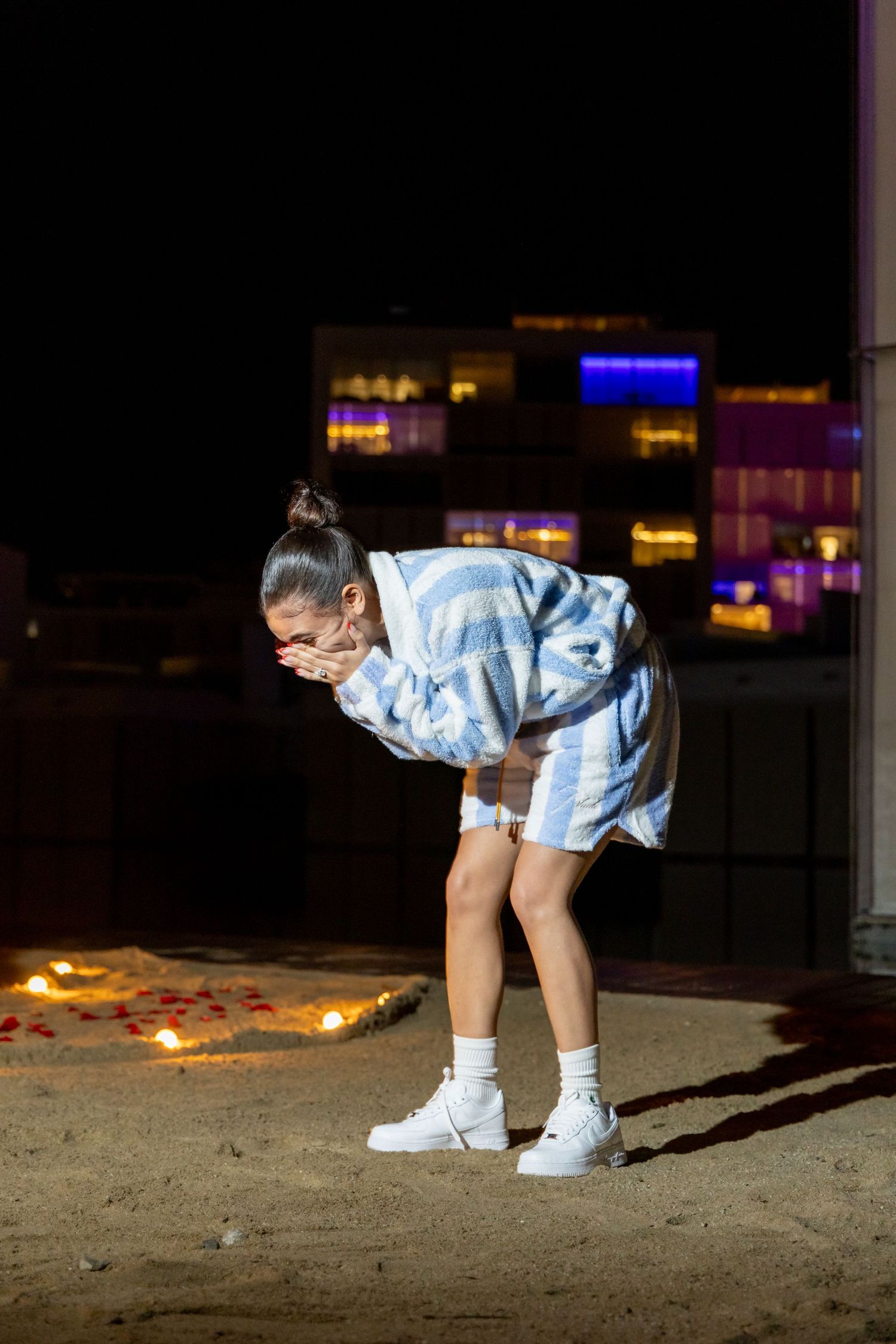 Person in a blue and white tie-dye outfit and white boots bends forward on a rooftop at night with city lights in background.