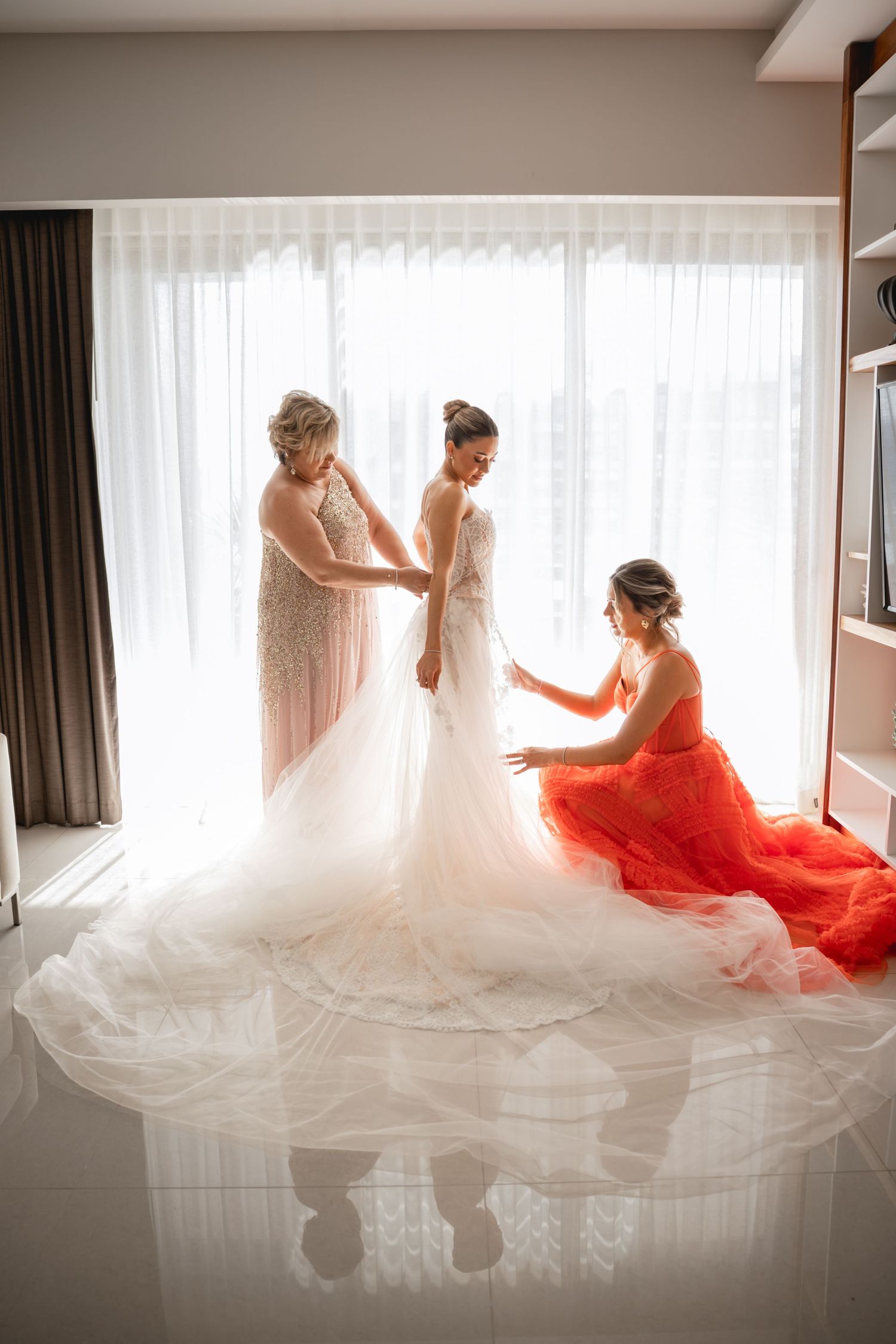 Bride in white dress with bridesmaid in red preparing in sunlit room.