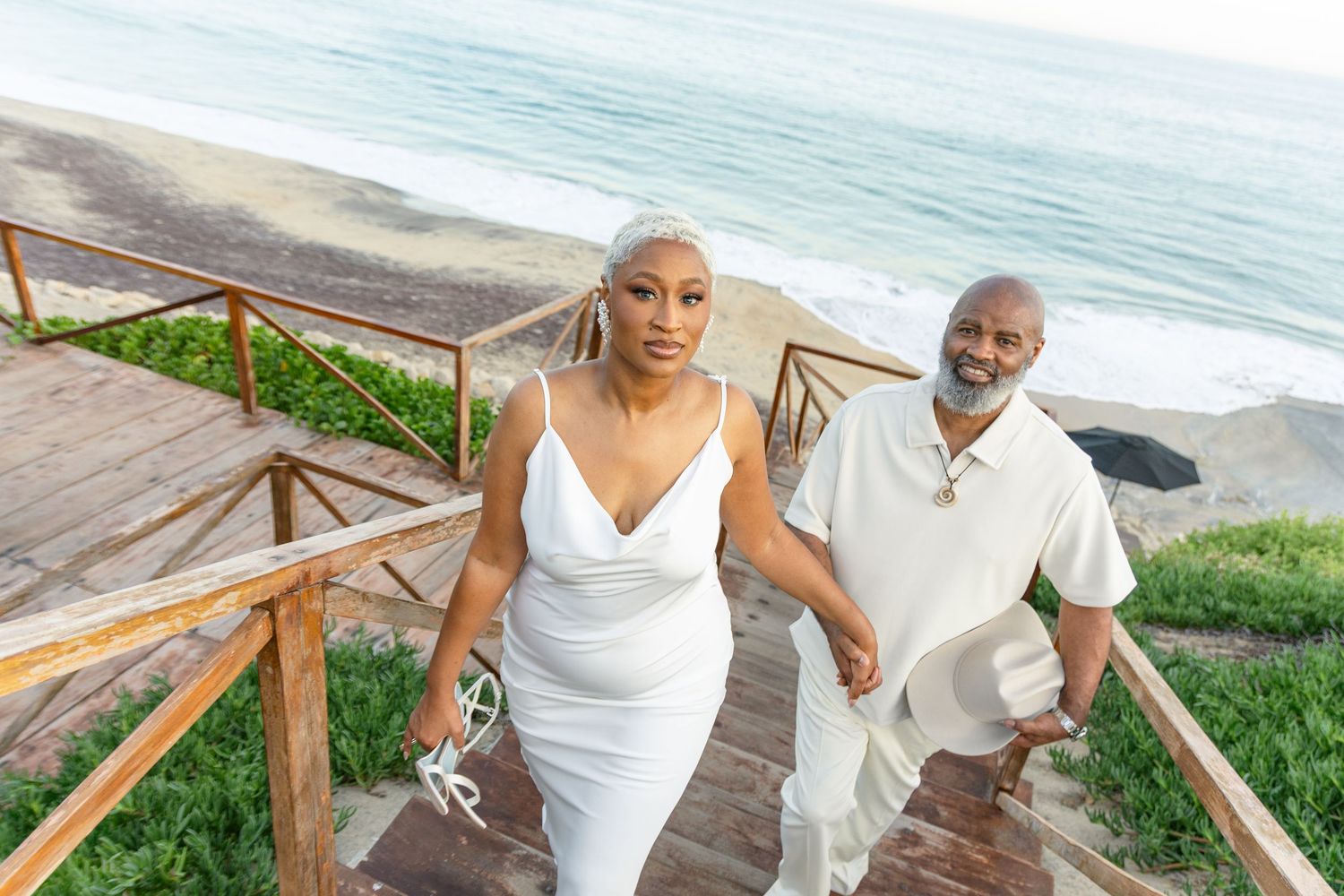 A beachside wedding celebration with a couple in white outfits walking along a wooden boardwalk.
