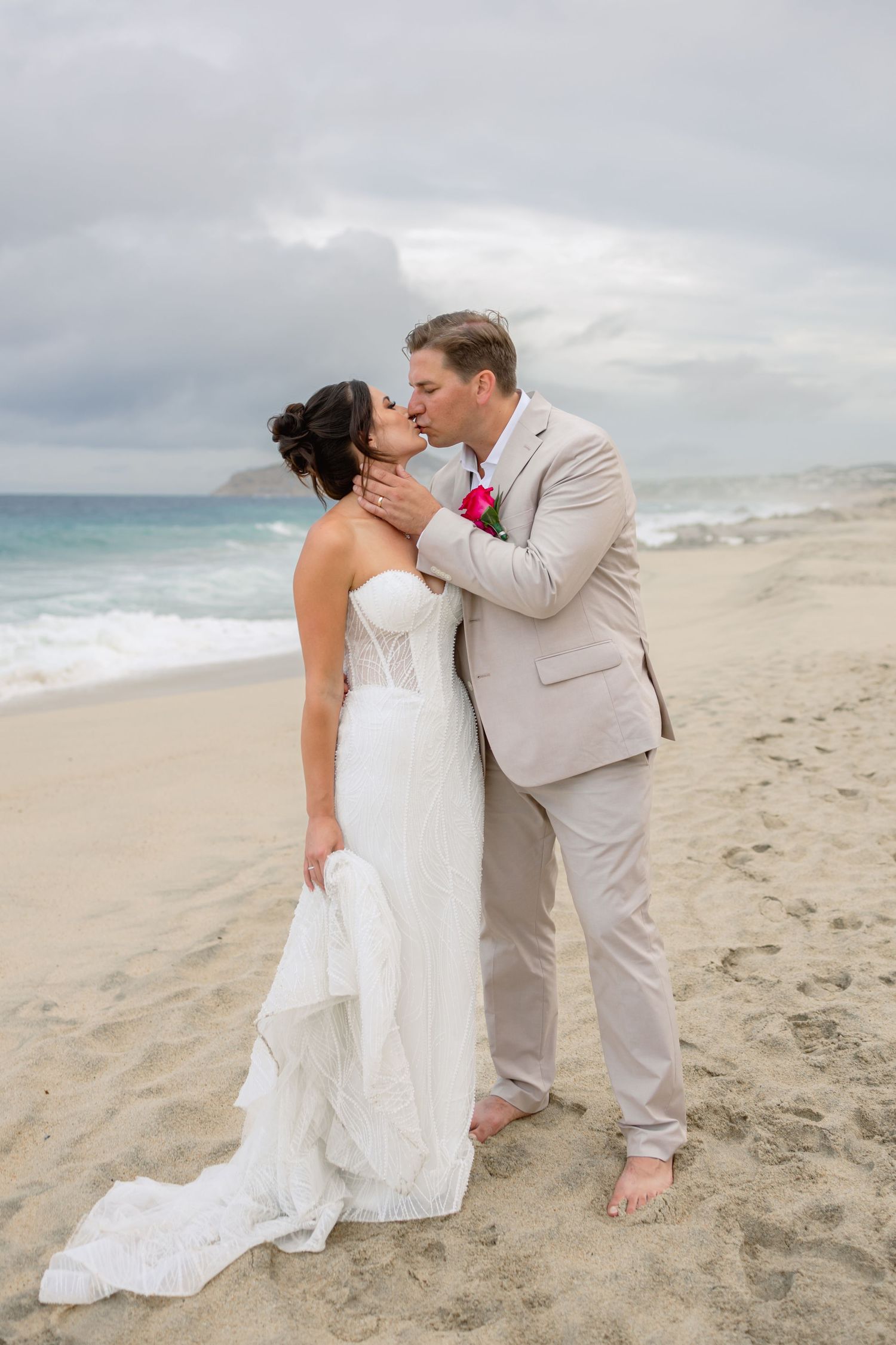 Romantic beach wedding kiss with waves and cloudy sky in background.