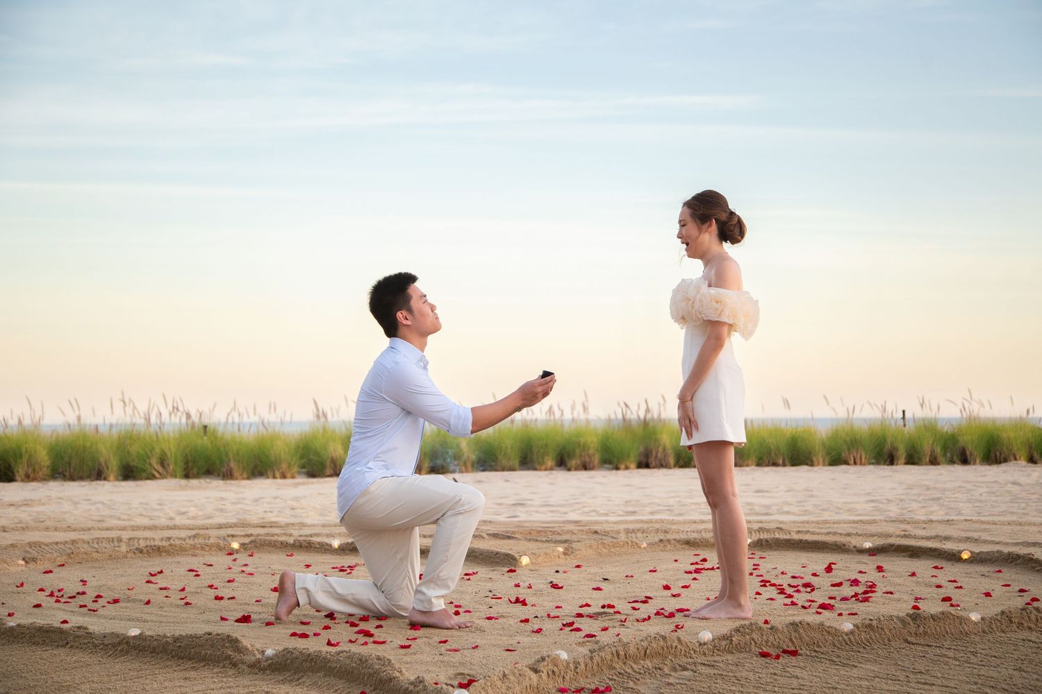 A romantic marriage proposal scene on a beach at sunset with rose petals arranged in a heart shape on the sand.