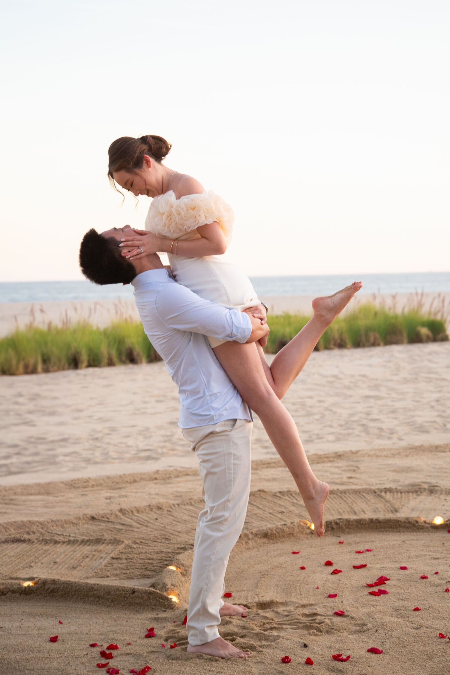 Romantic couple embracing on beach at sunset with rose petals scattered on sand.