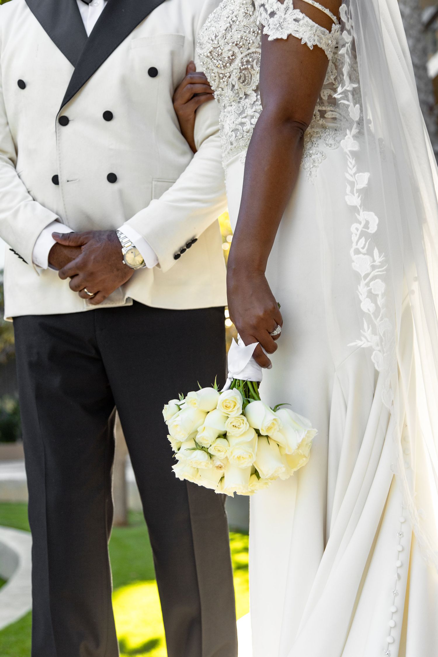 Detail shot of couple in white wedding attire holding white rose bouquet during outdoor ceremony.