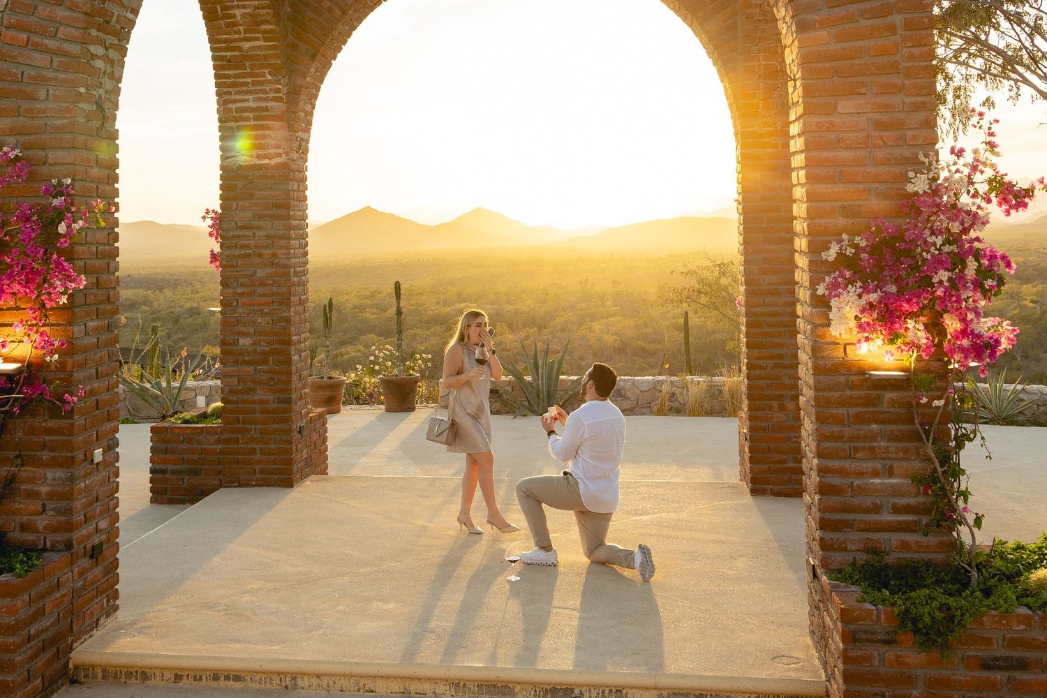 A silhouetted marriage proposal at sunset with mountains in the background, framed by brick columns.