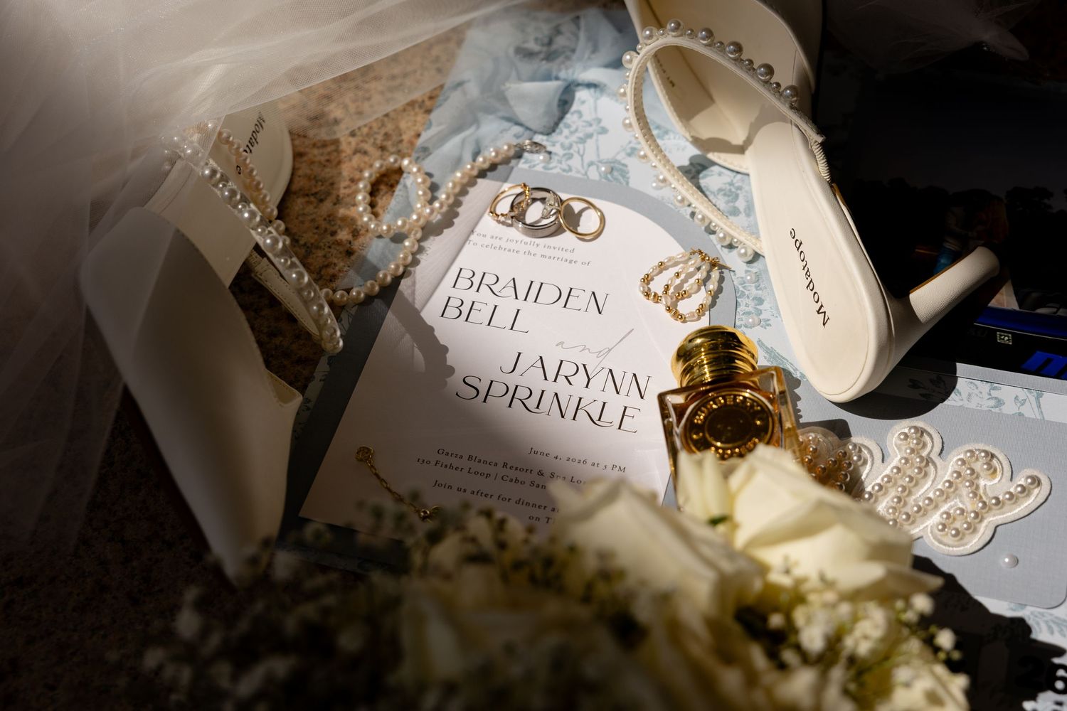 Wedding flat lay featuring invitation for Braiden Bell and Laryna Sprinkle with gold rings, pearl necklace, and bridal accessories.