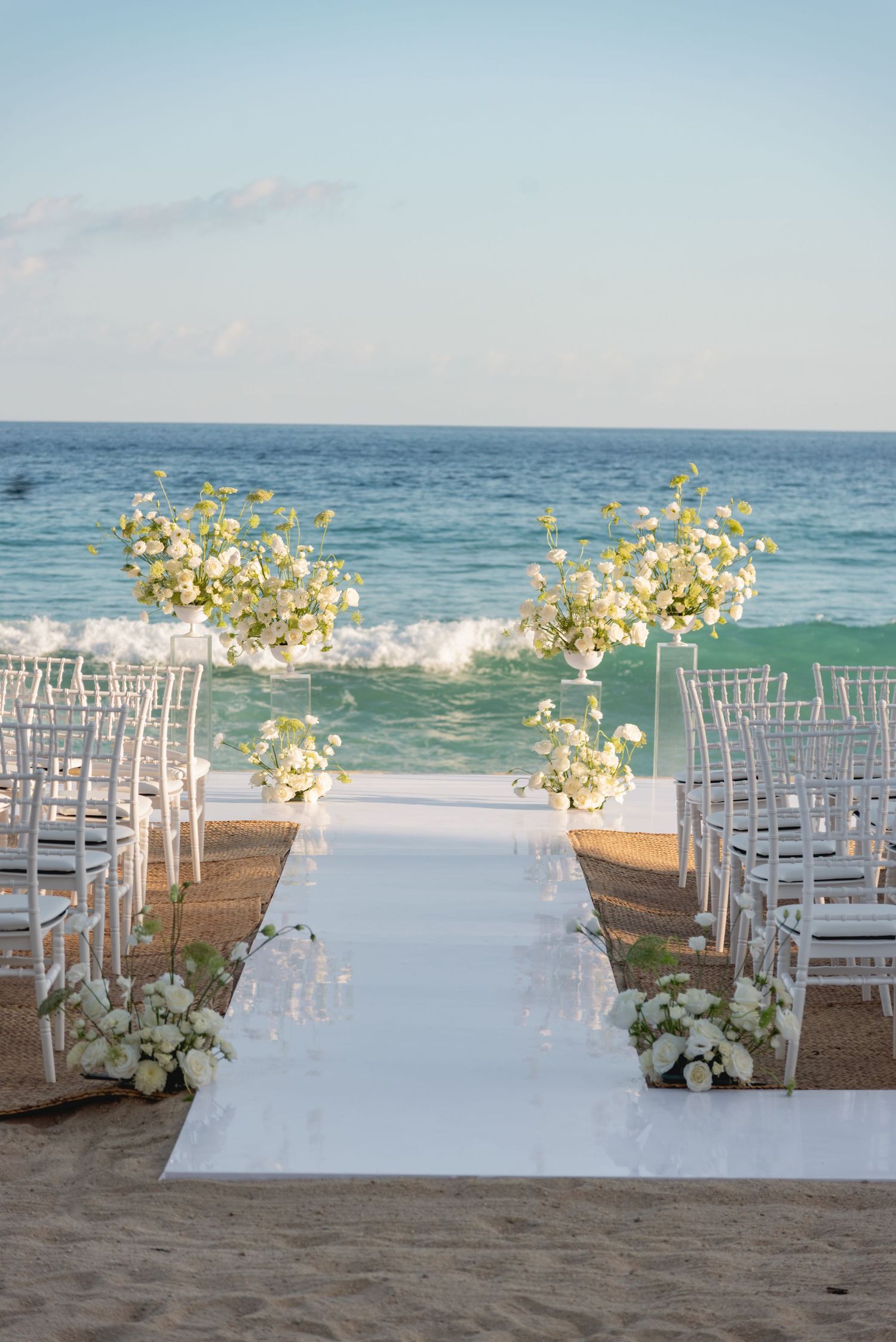 Beach wedding ceremony setup with white chairs and floral arrangements along the ocean.