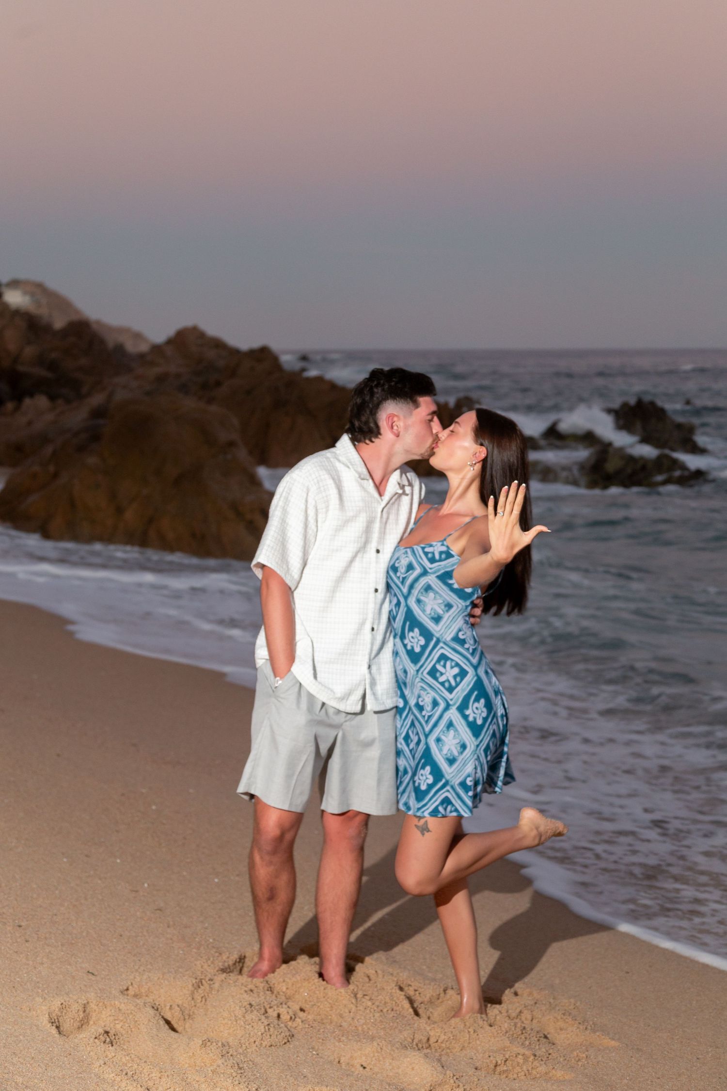 A couple shares a romantic kiss on a sandy beach at sunset with ocean waves and rocky coastline in the background.