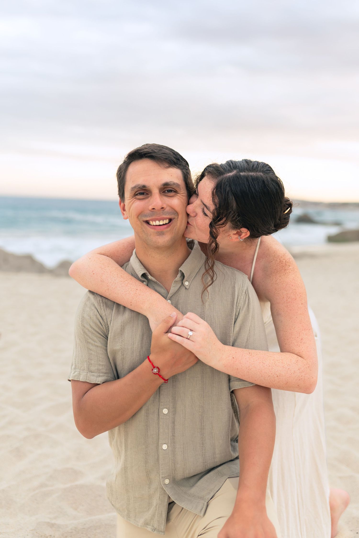 A happy couple embraces on a beach at sunset with ocean waves and sand in the background.