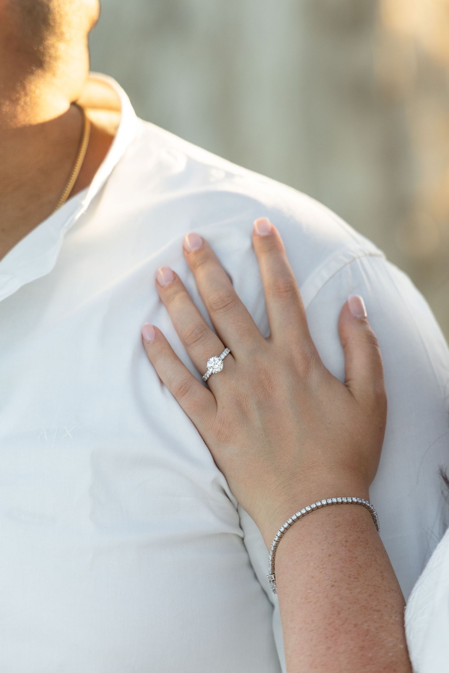 A close-up view of hands wearing a diamond engagement ring and bracelet against a white fabric background.