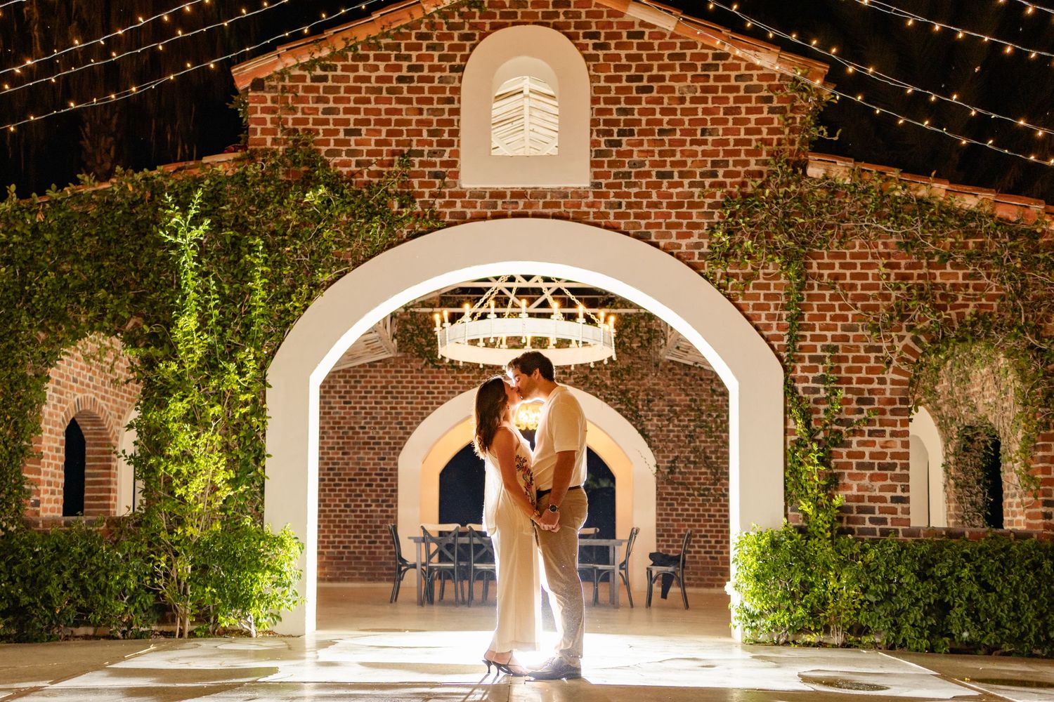 String lights illuminate a rustic brick archway walkway with climbing vines and garden landscaping at dusk.