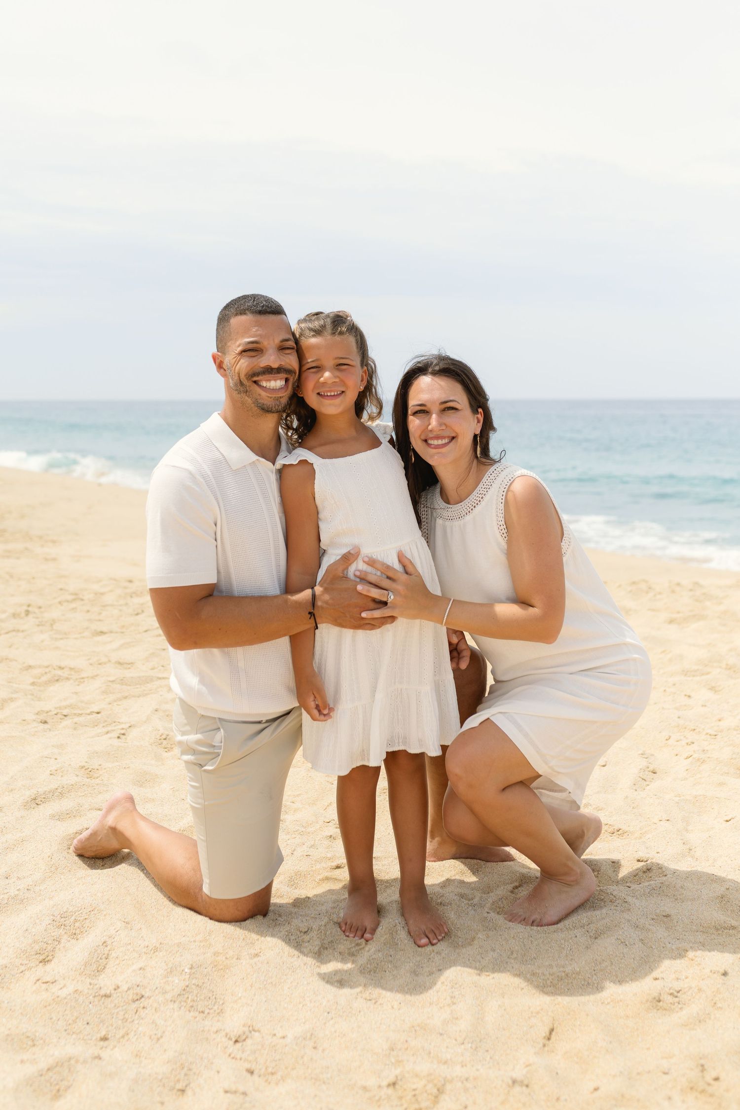 A family wearing white outfits poses together on a sunny beach with ocean waves in the background at diamante Cabo San Lucas