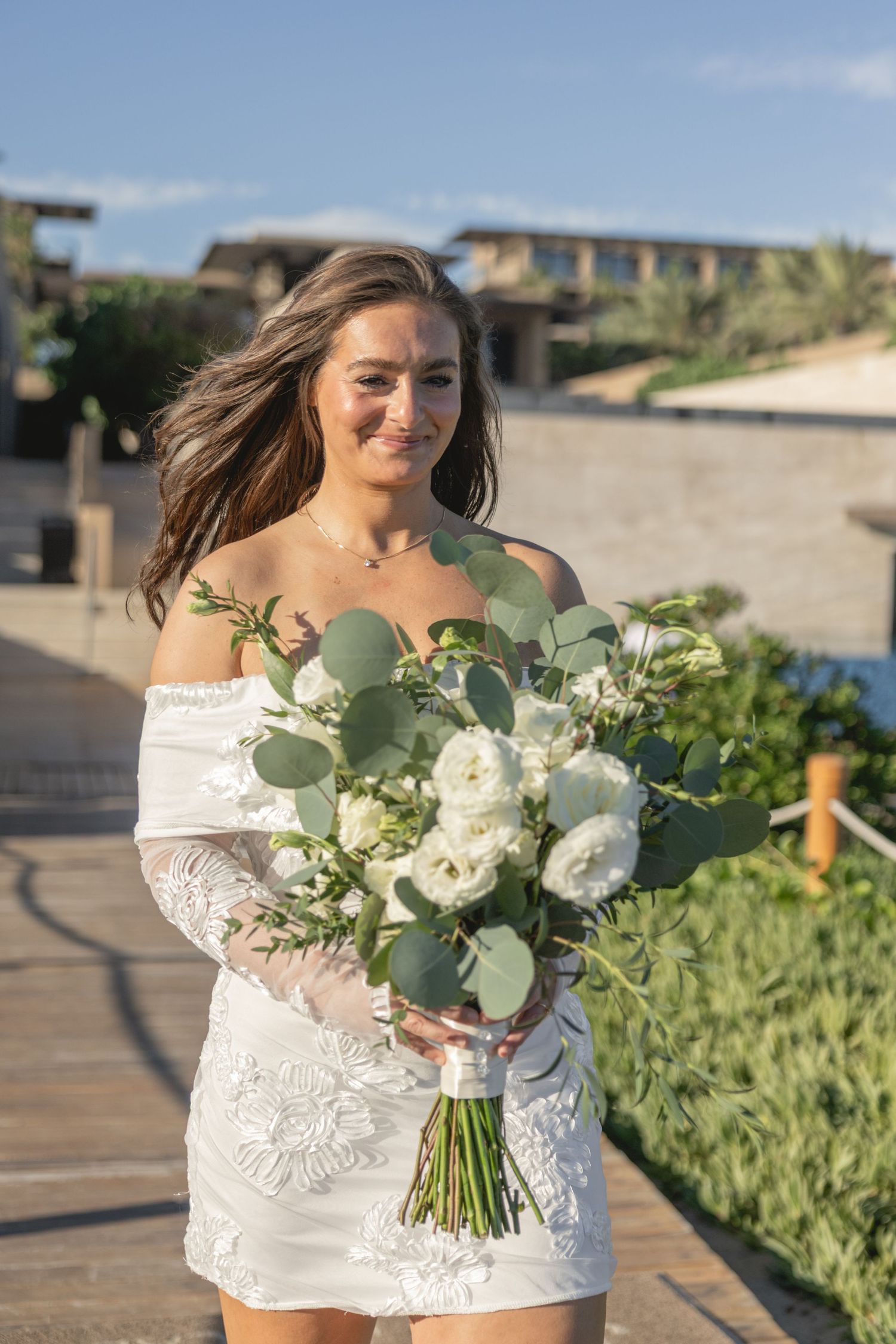 A person in white lace holds a stunning bouquet of white flowers and eucalyptus against an ocean backdrop.