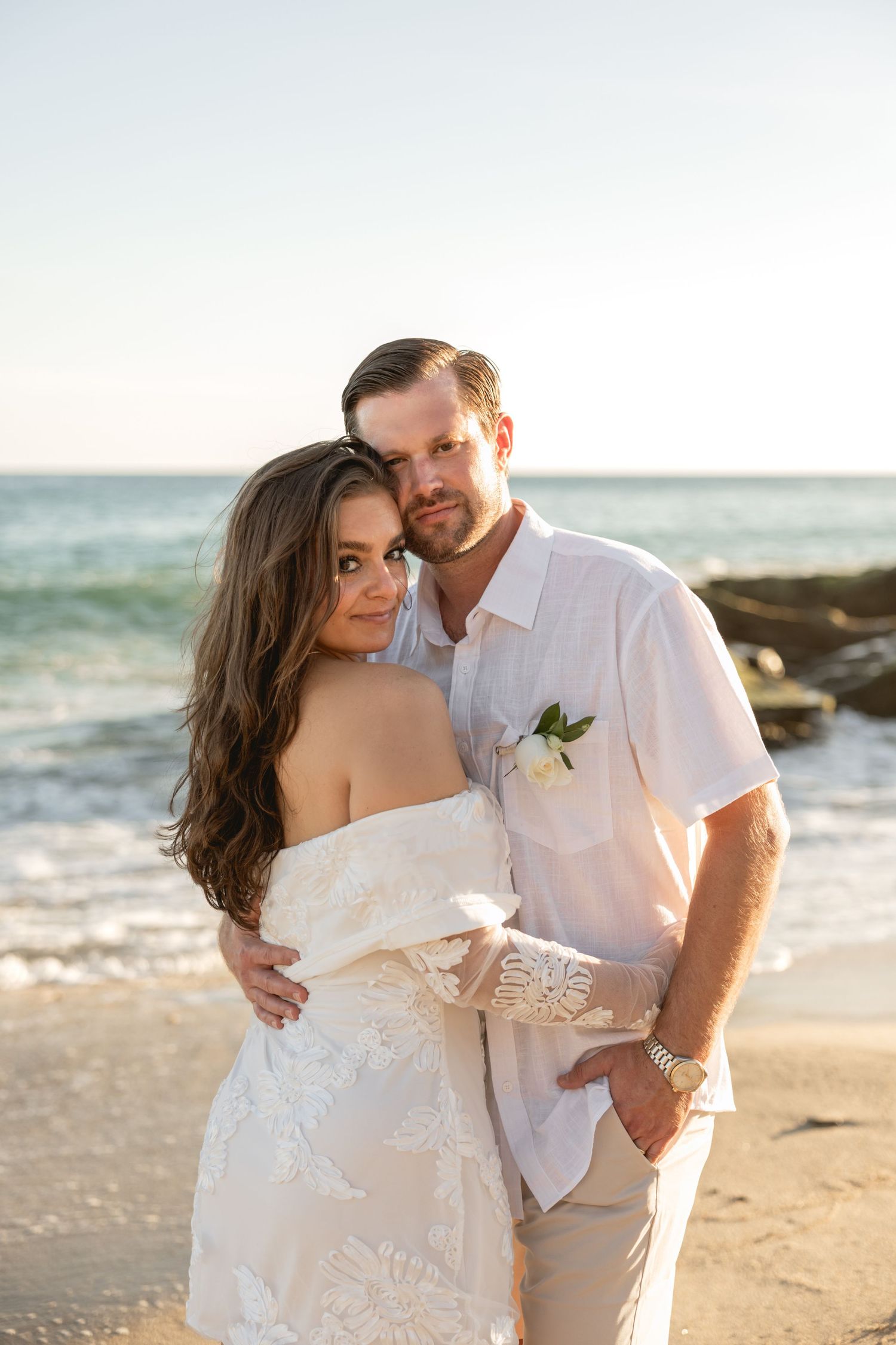 Bride and groom embrace on a beach at sunset, wearing white wedding attire with ocean waves in the background.