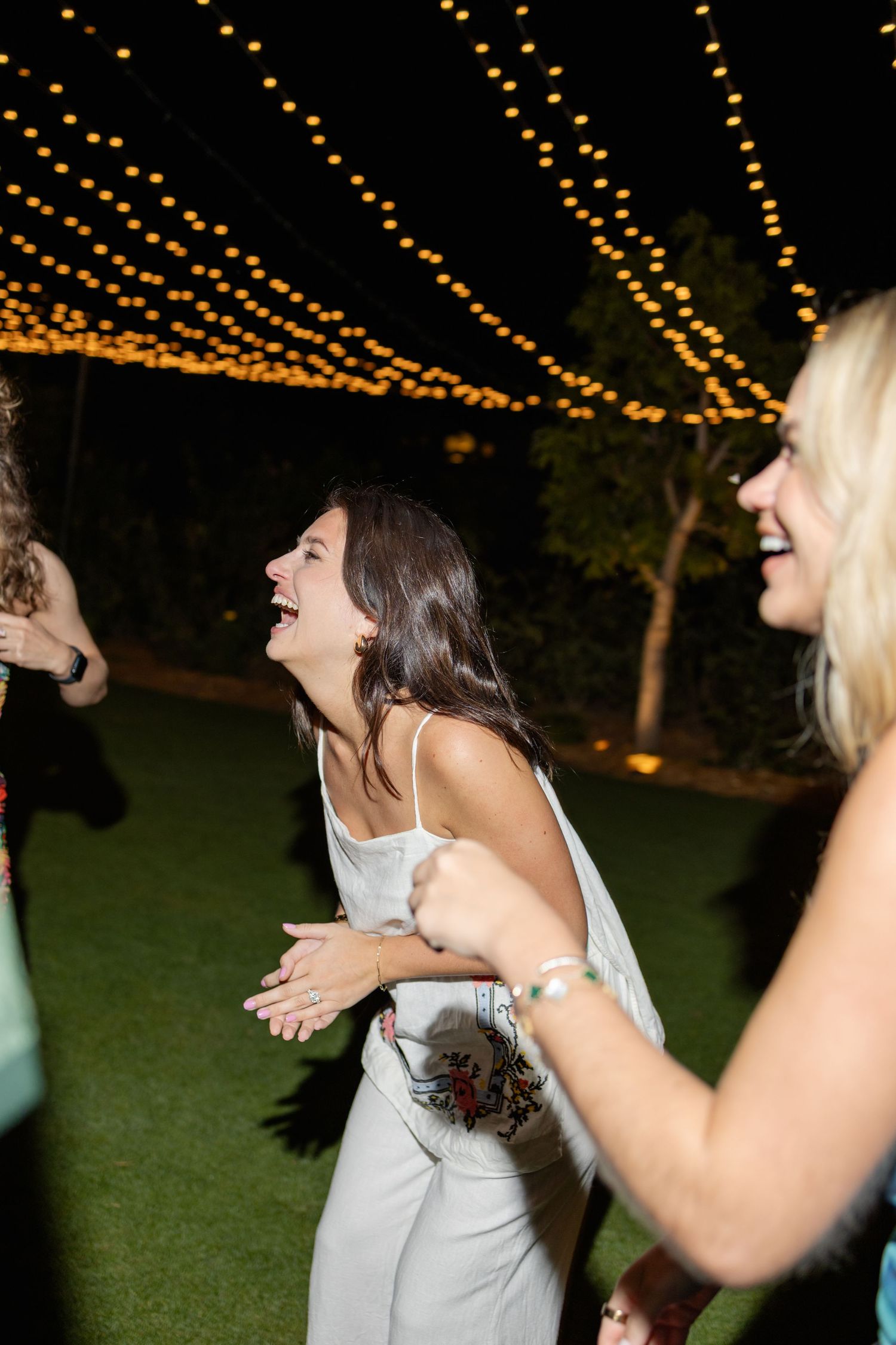 A group of people laughing together outdoors at night under strings of glowing lights.