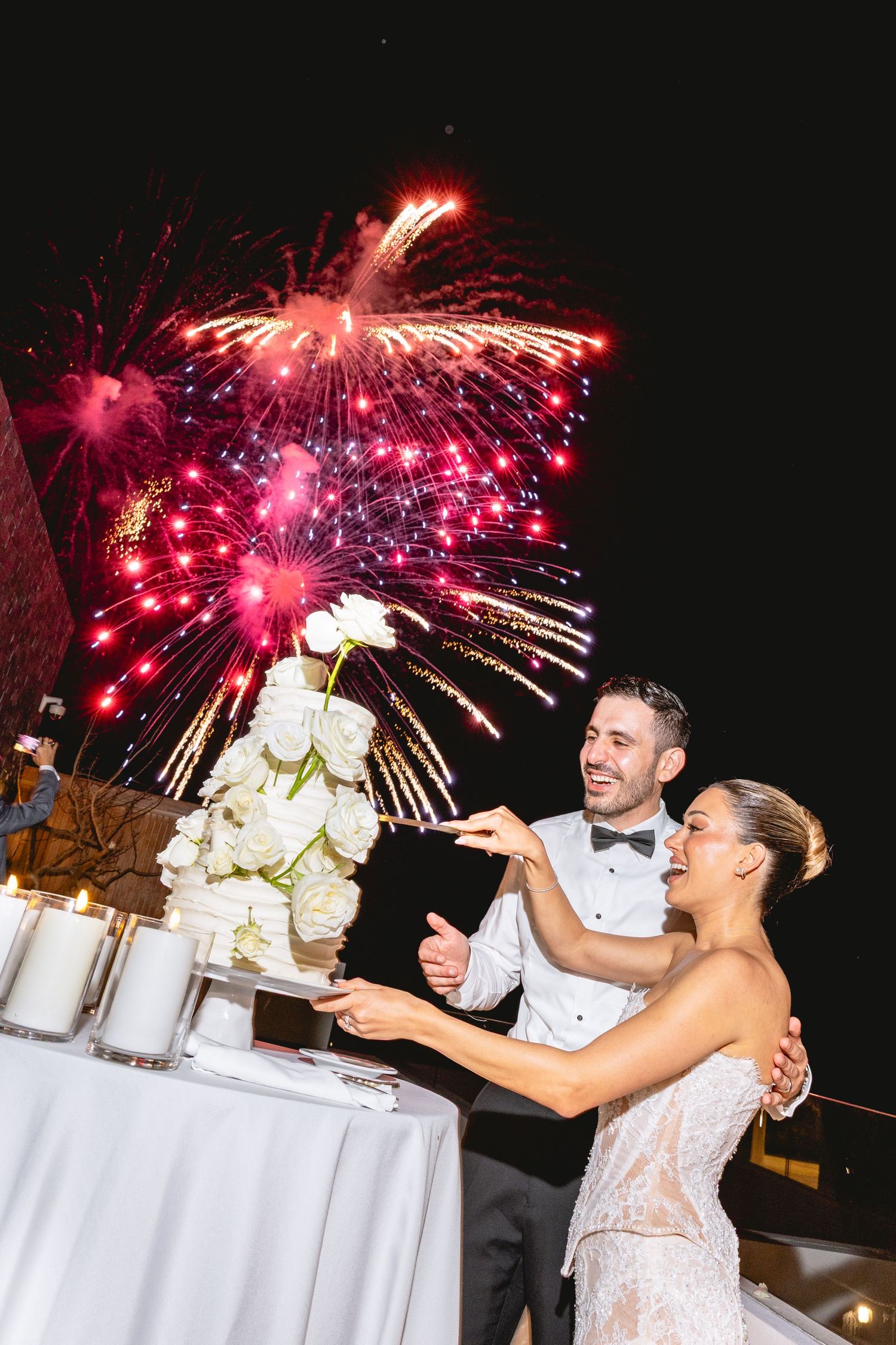 Vibrant pink fireworks explode behind a wedding reception table with white floral centerpieces.