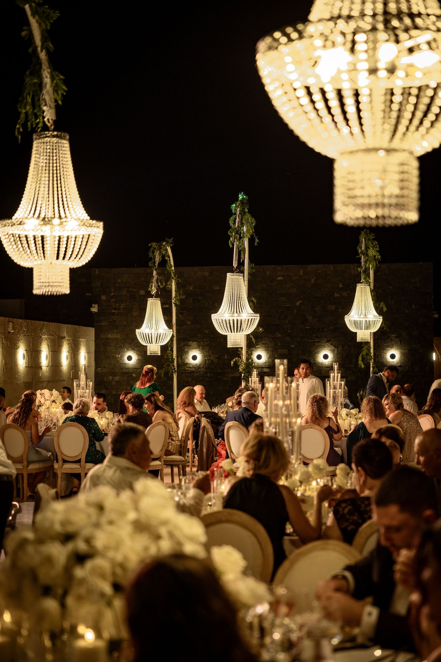 Crystal chandeliers illuminate an evening wedding reception with guests seated at elegant tables.