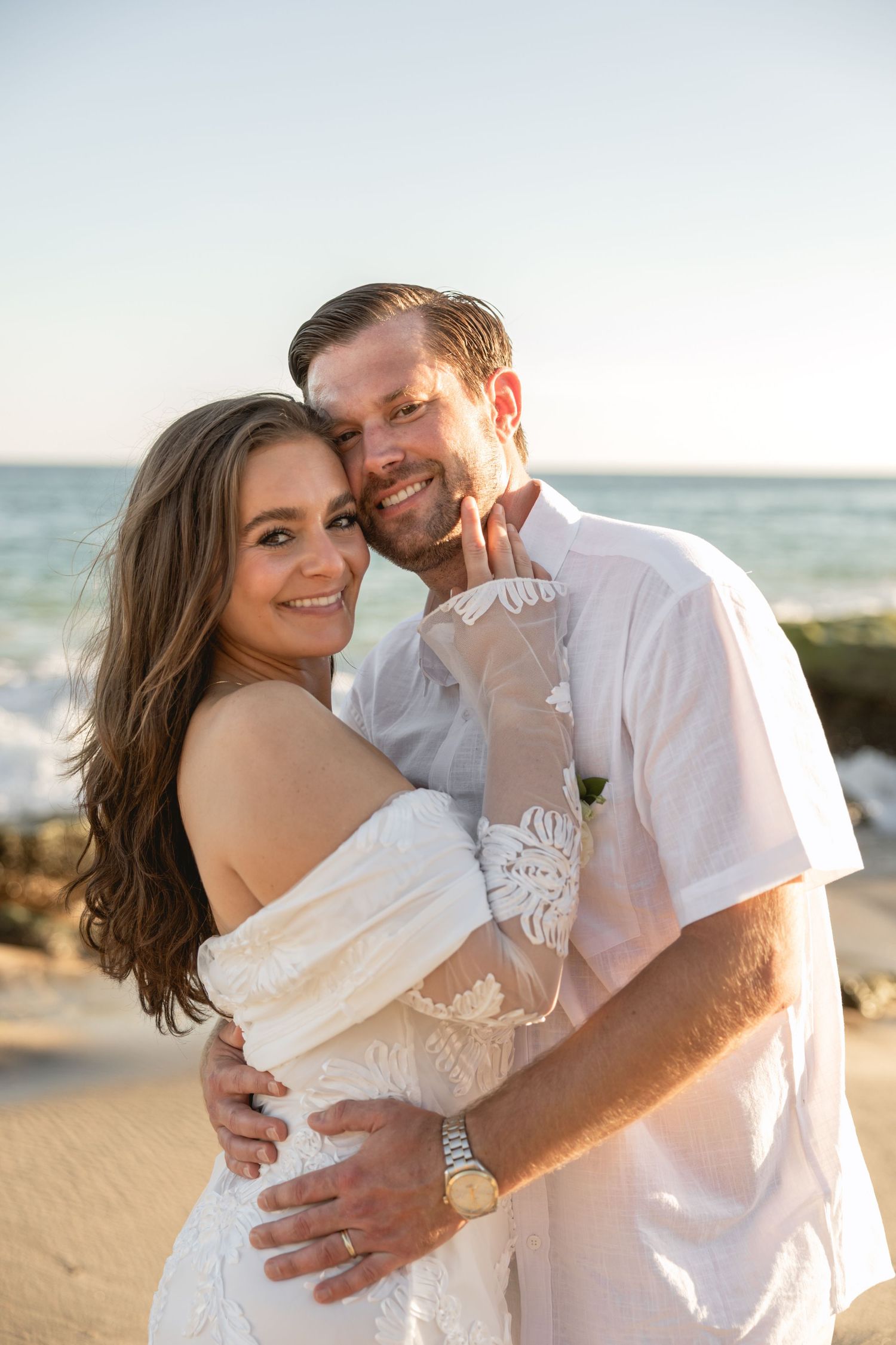 A romantic beach portrait of a couple embracing in white attire against an ocean backdrop.