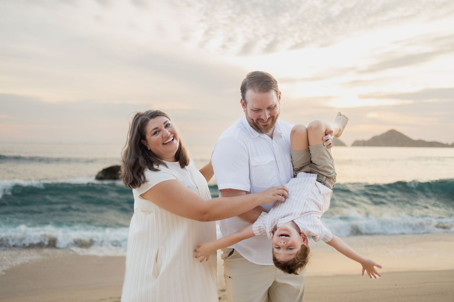 Happy family of three playing on a sandy beach at sunset, parents laughing as they swing their child upside down.