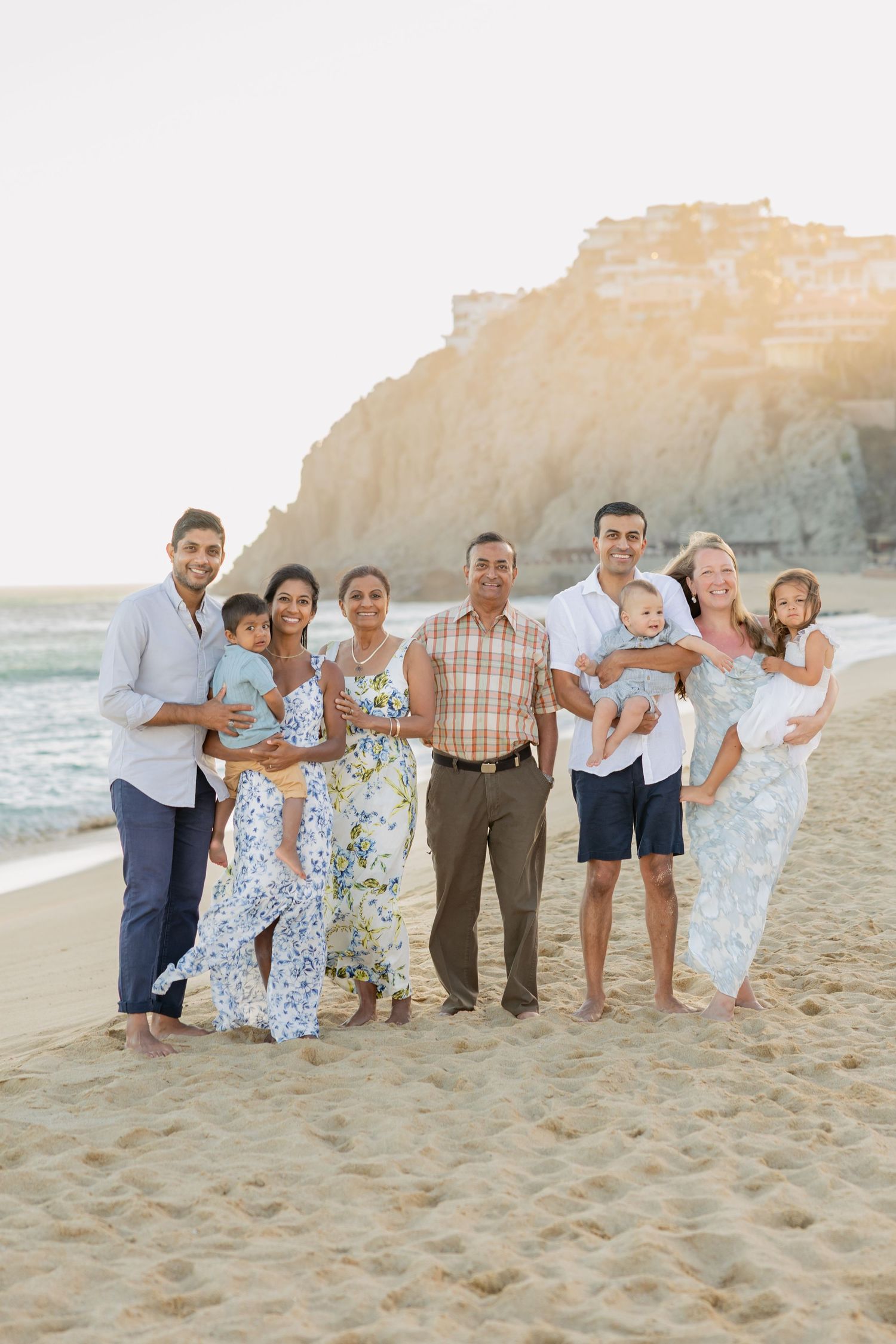 Multi-generational family portrait on sandy beach at golden hour with rocky cliffs in background.