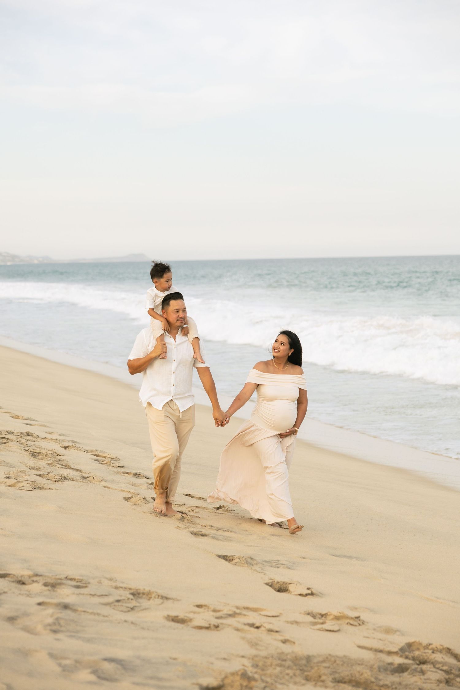 Pregnant woman in white gown walks on beach with partner and child on shoulders, family maternity photo session.