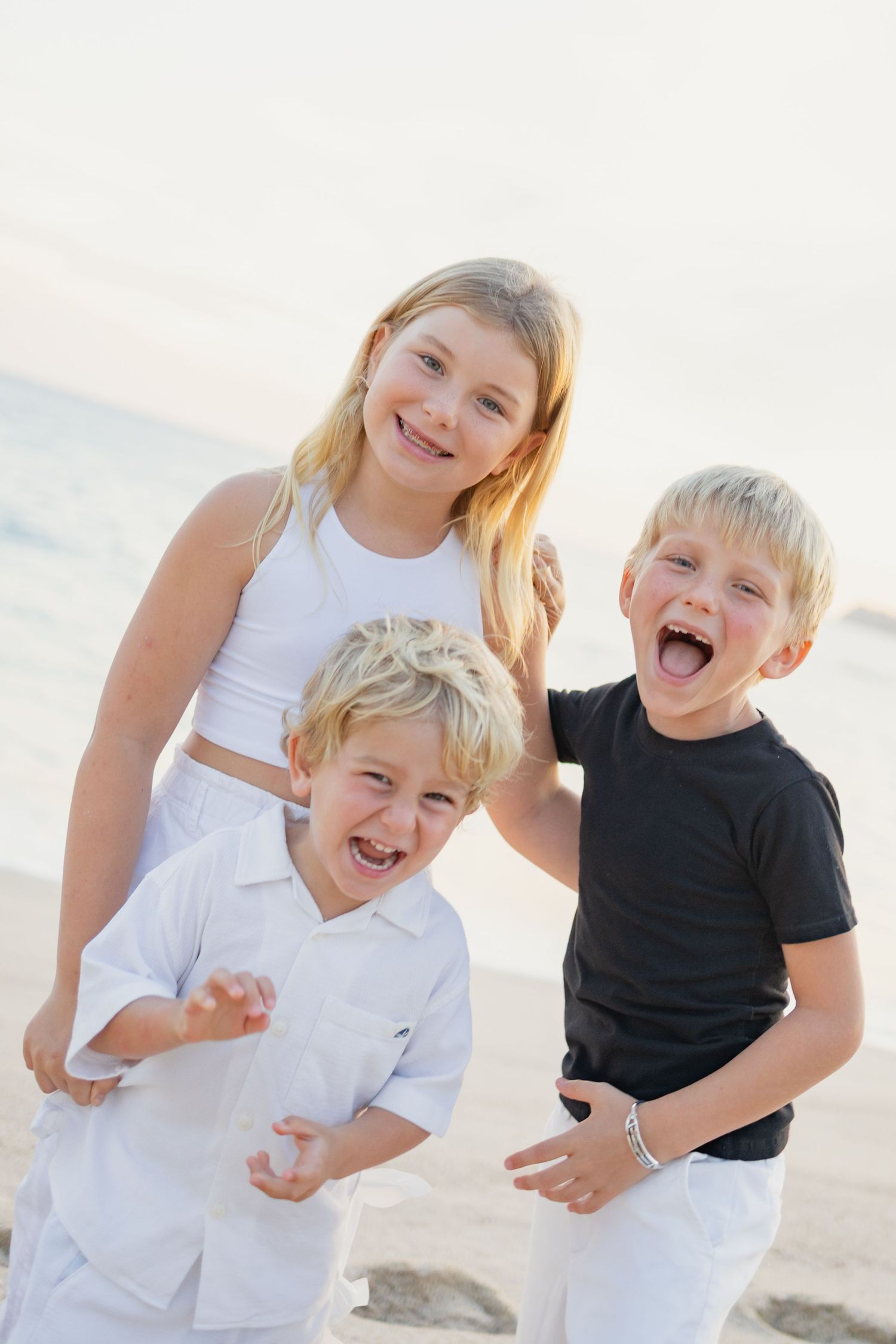 Children playing and laughing together on a sunny beach in white and black outfits.