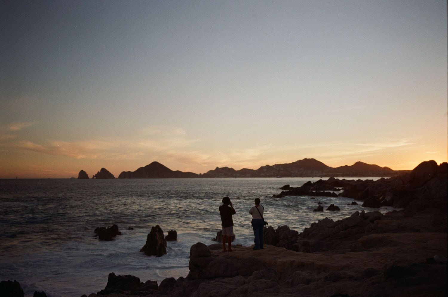 Silhouetted people stand on rocky shore watching a stunning ocean sunset with mountainous rock formations in Cabo San Lucas.