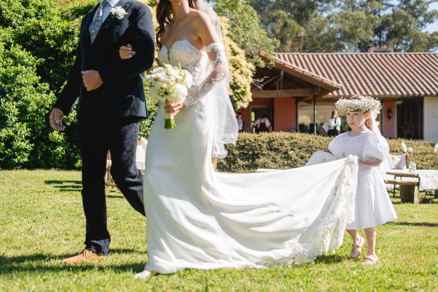 Wedding dress train being held on sunny lawn with Spanish-style building in background.