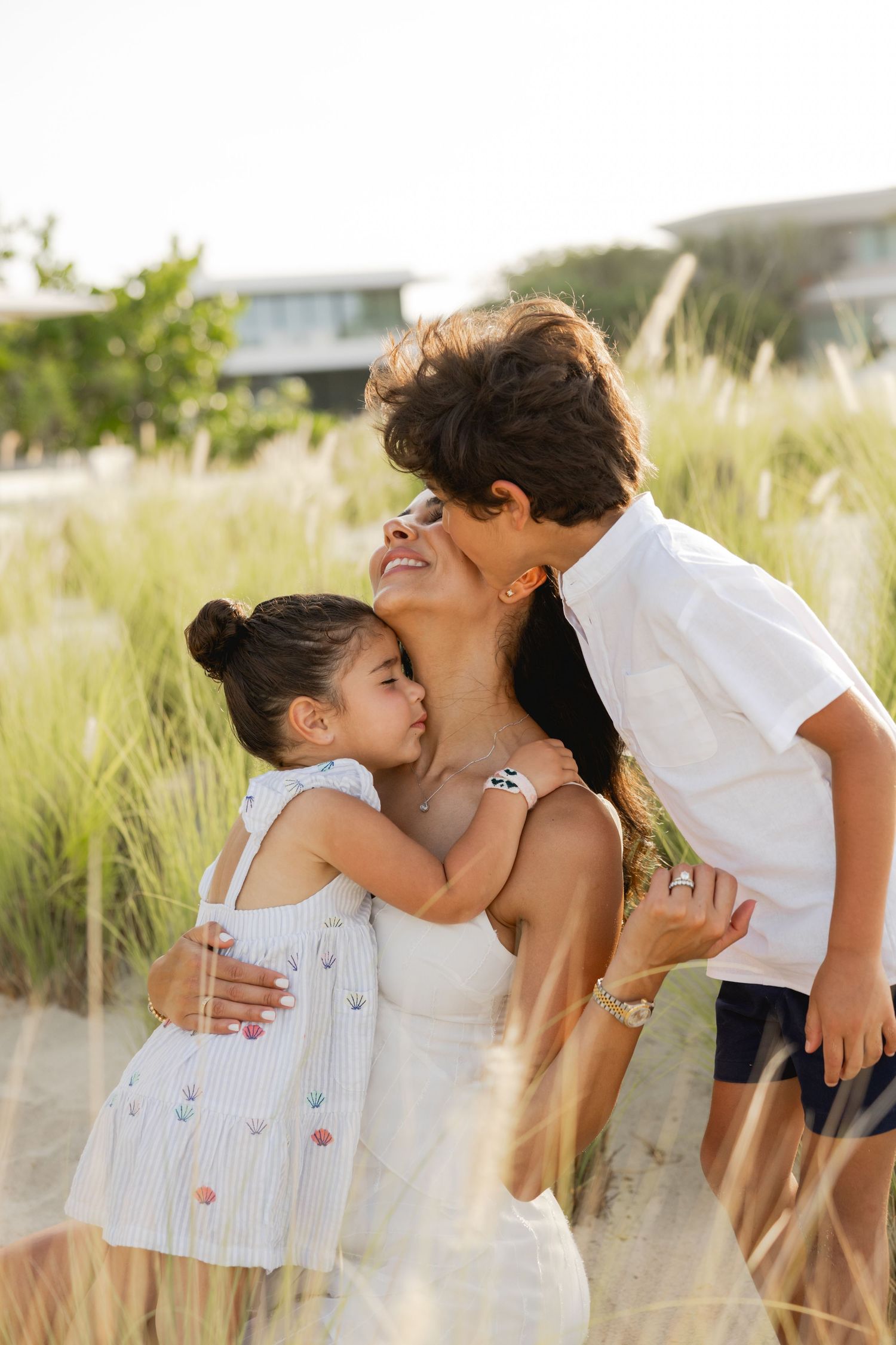 A mother smiles as her two young children embrace her outdoors near tall grass on a sunny day.