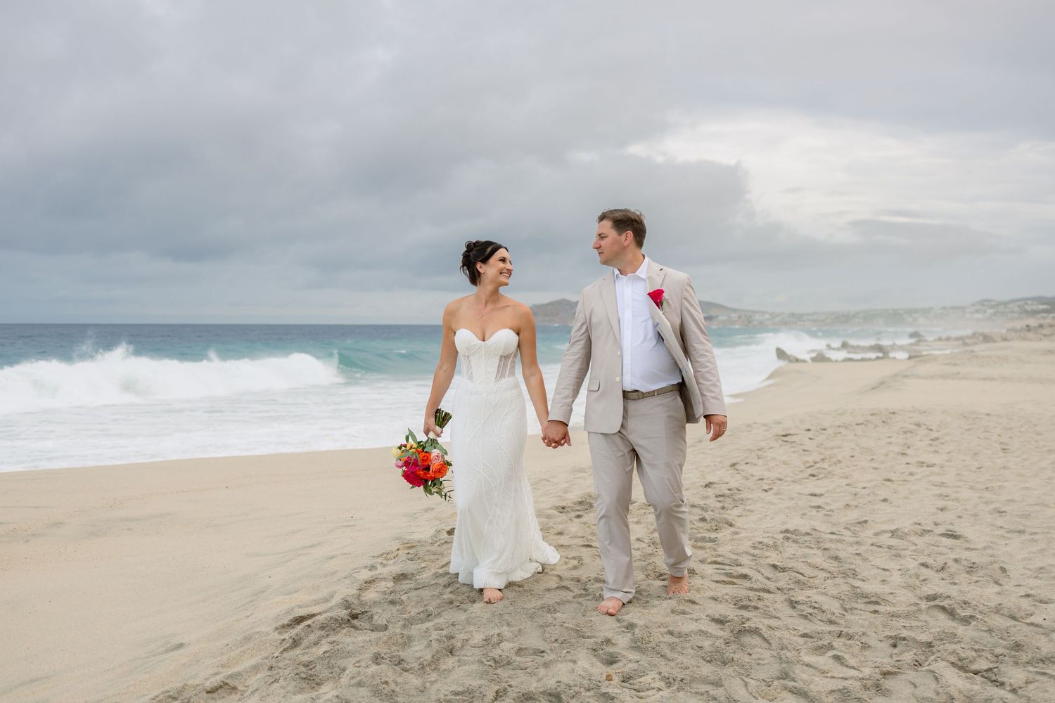 Newlyweds walking hand in hand along a scenic beach with cloudy skies and ocean waves.