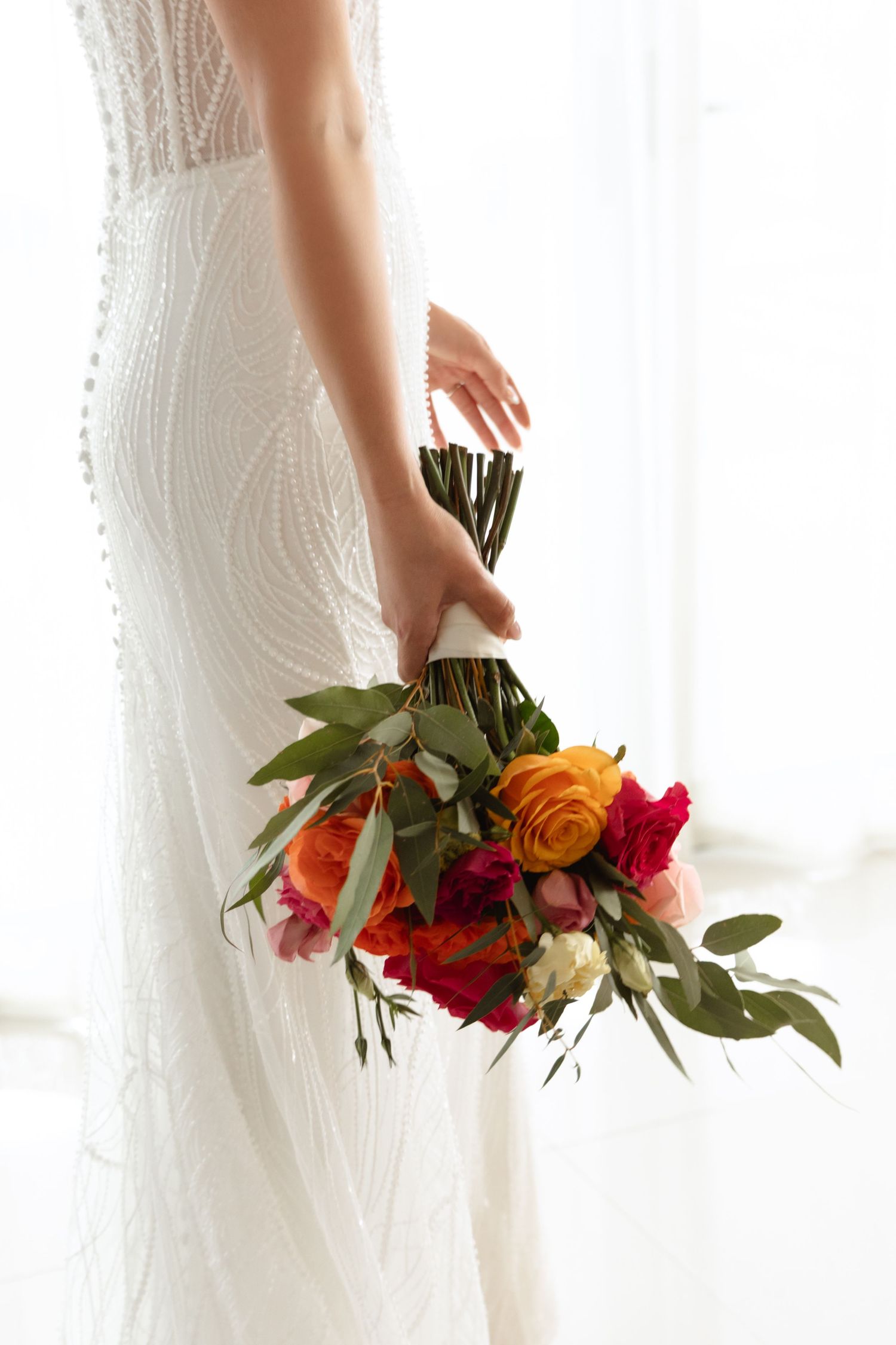 Elegant wedding dress detail with colorful bouquet featuring orange and red flowers.