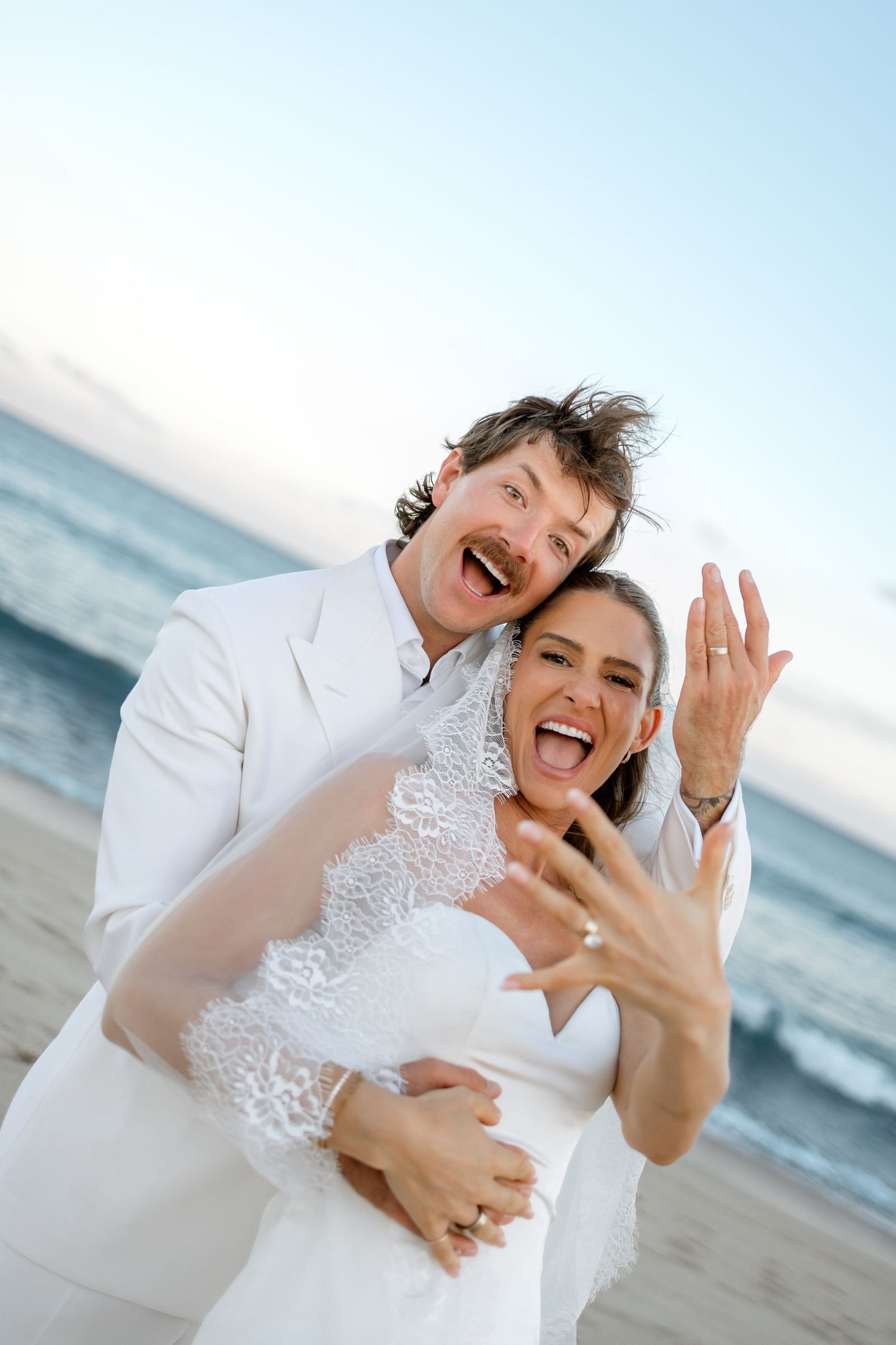 Joyful newlyweds in white attire showing wedding rings on a beach, laughing and embracing by the ocean.