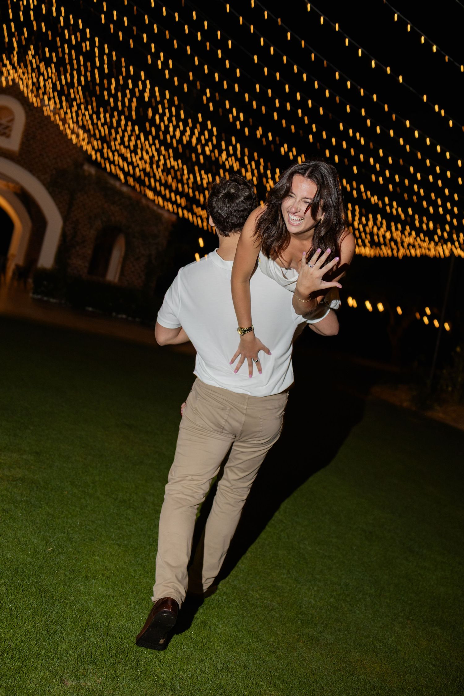 A person dances under a canopy of warm glowing string lights on a grassy lawn at night.