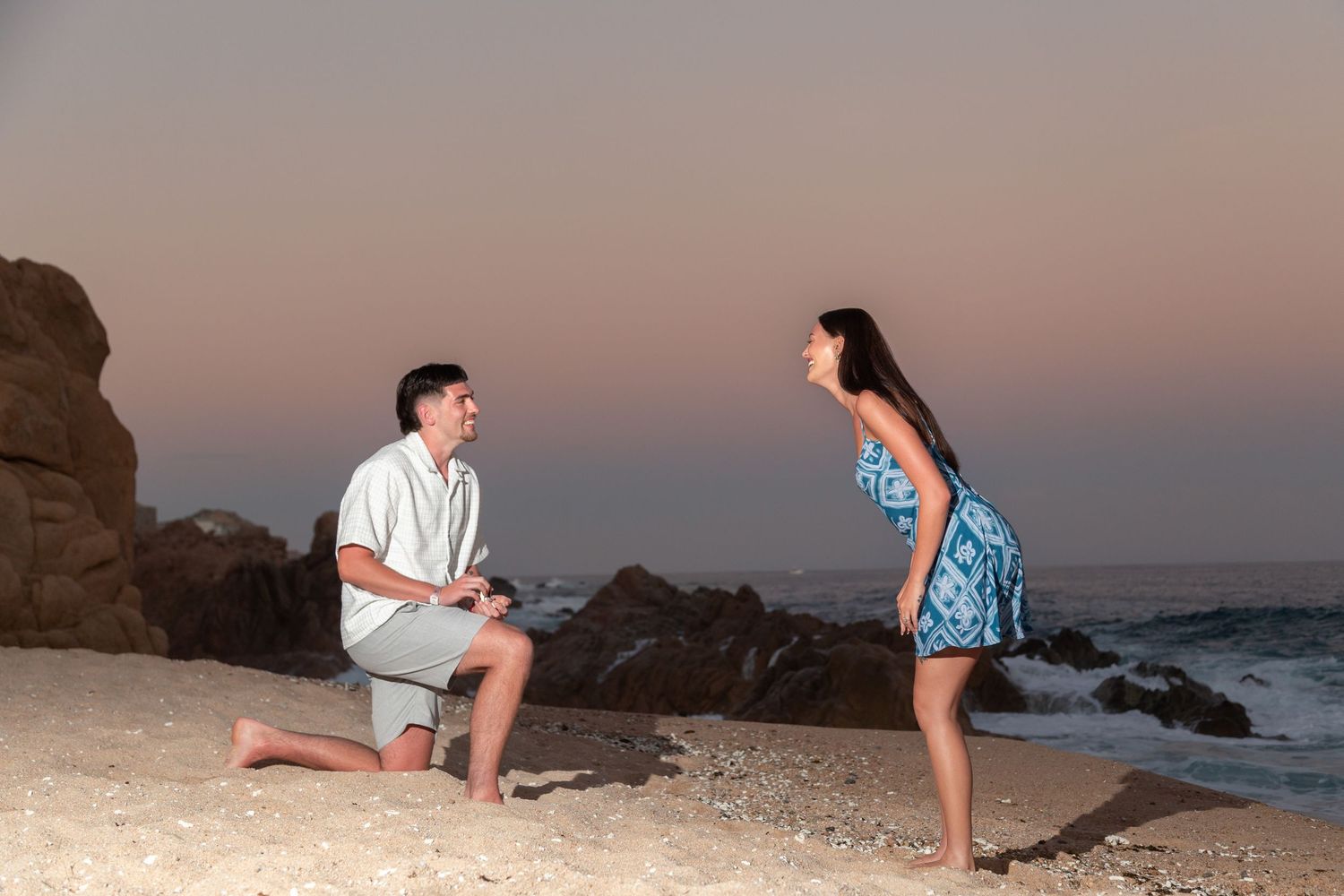 A romantic sunset beach proposal scene with two people on the sand near ocean waves and rocky cliffs.