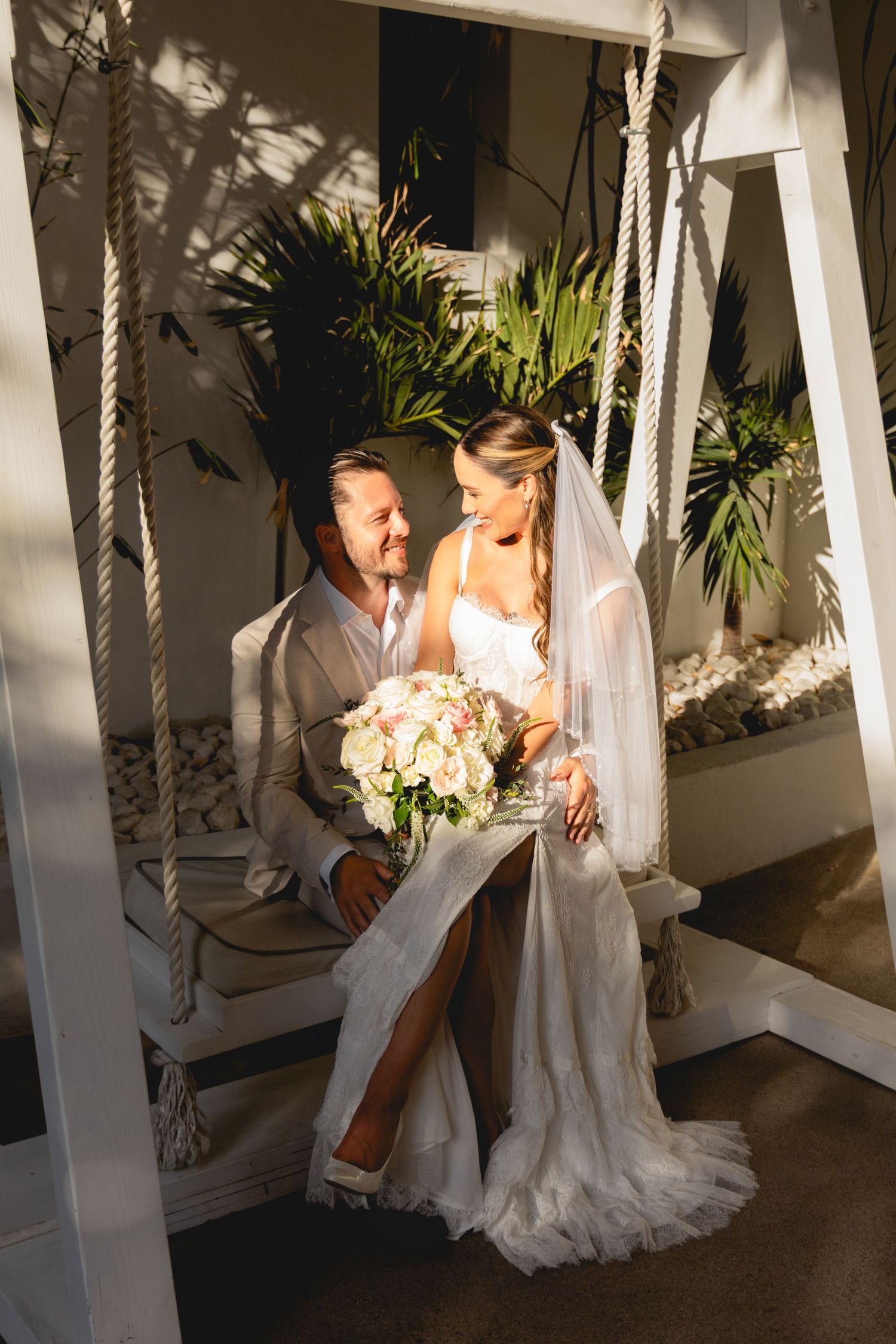 Couple shares tender moment on white rope swing with tropical garden backdrop.