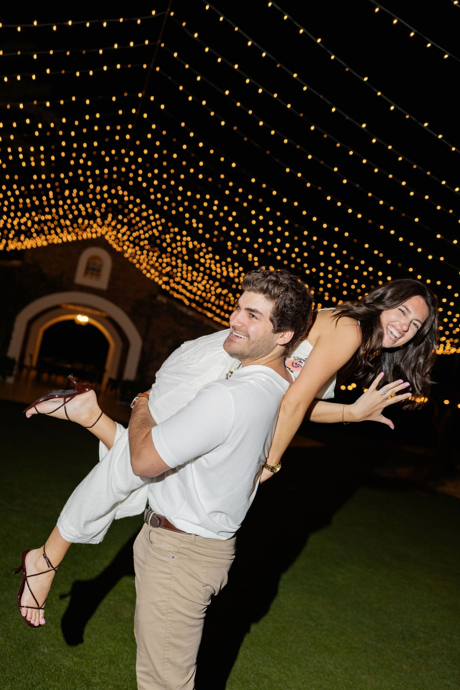 A couple dances under a canopy of twinkling string lights at an outdoor evening event.
