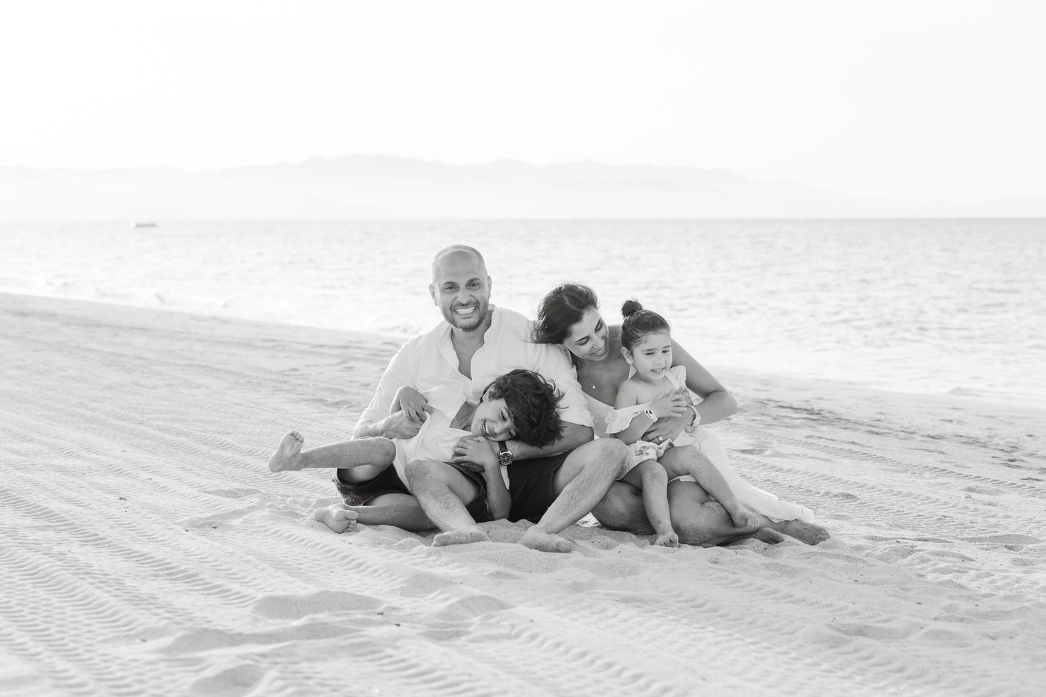 Black and white beach photo of a smiling man sitting on sand with three children climbing on him near the ocean.