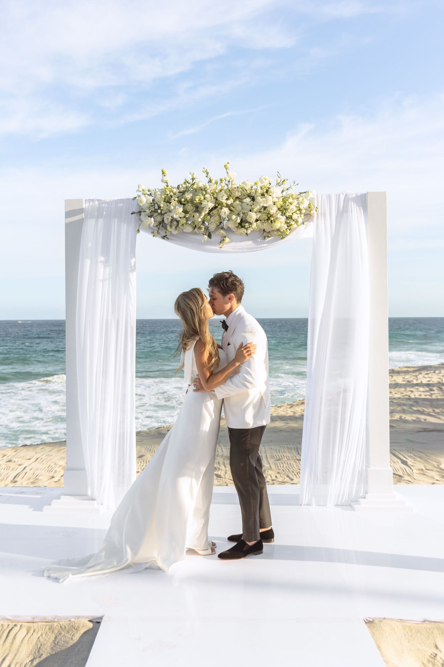 Romantic beach wedding ceremony with white draped canopy and ocean waves in background.