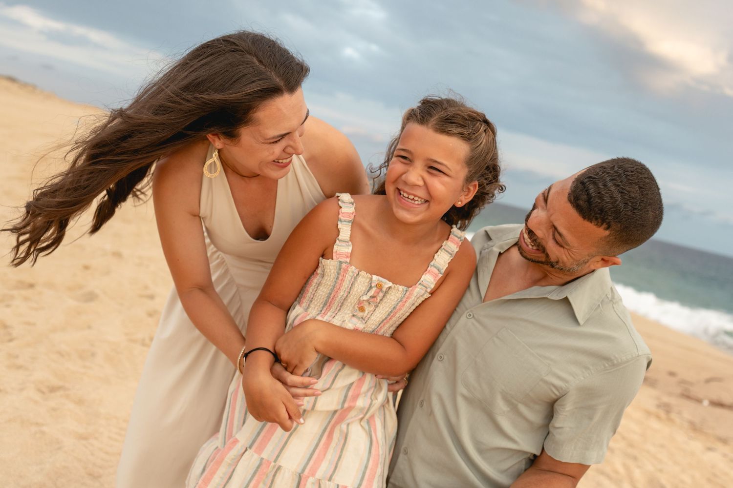 Happy family of three laughing together on a sandy beach, with ocean waves in the background.