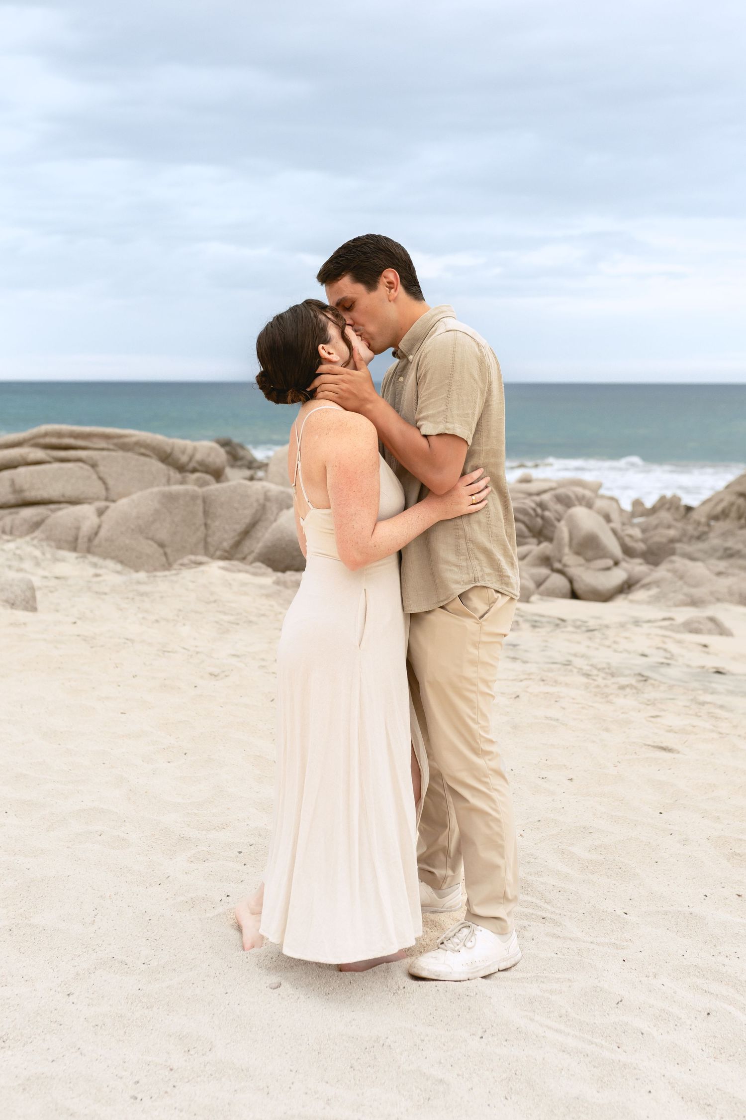 A couple shares a romantic kiss on a sandy beach with ocean waves and rocky outcrops in the background.