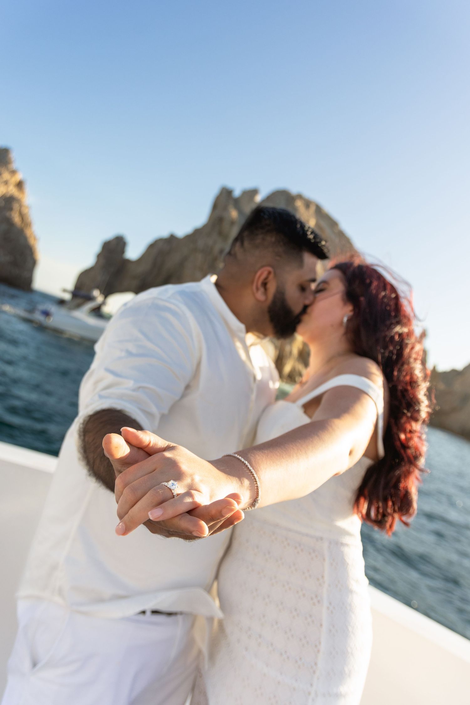 A romantic couple in white outfits share intimate kisses on a beach with ocean rocks in the background.