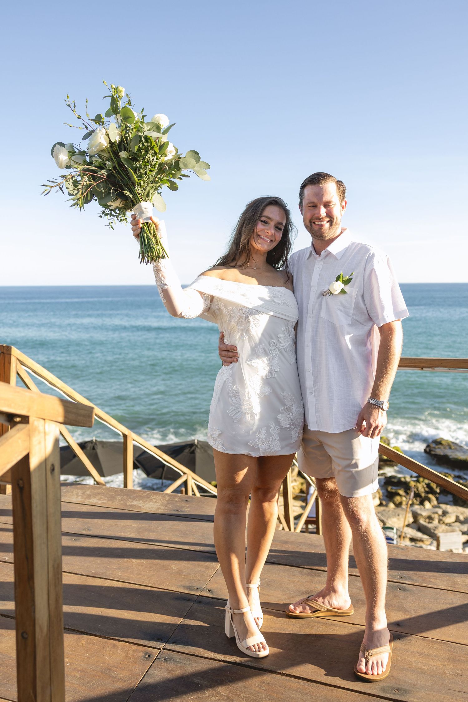 Couple in white attire celebrating on oceanfront deck with bouquet.